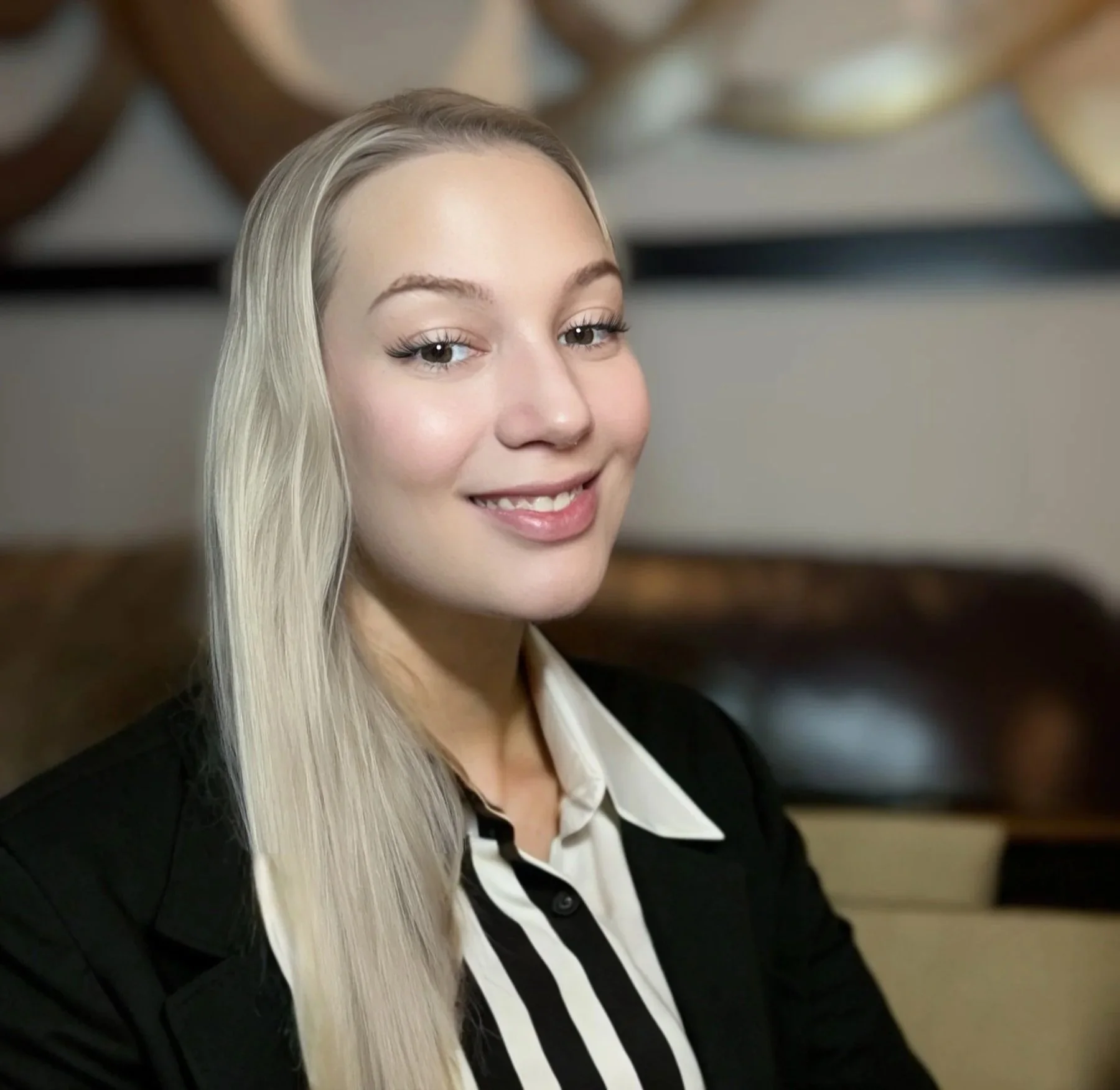 A young woman with long blonde hair smiling at the camera, dressed in a black blazer and white shirt, sitting indoors with art on the wall behind her.