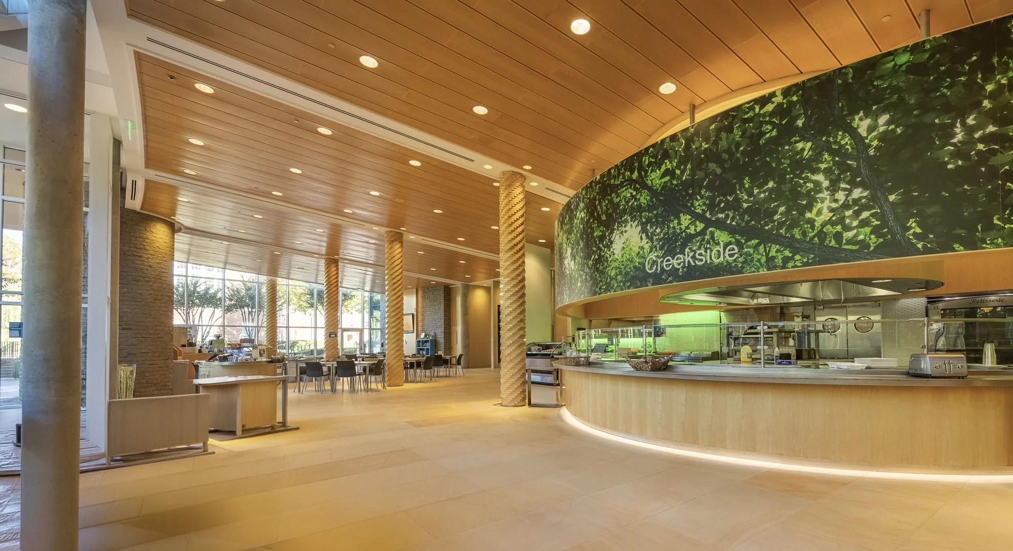 Interior of a spacious cafeteria with wooden ceilings, large windows, and a curved green wall with trees and the word "Creekside" on it. There are tables and chairs, and a food serving area.
