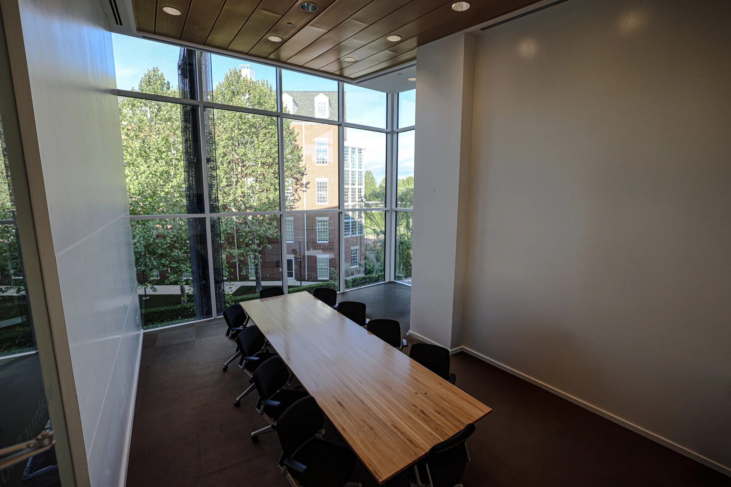 An empty conference room with a long wooden table and black chairs, large floor-to-ceiling windows showing trees and buildings outside, and a wooden ceiling with recessed lighting.