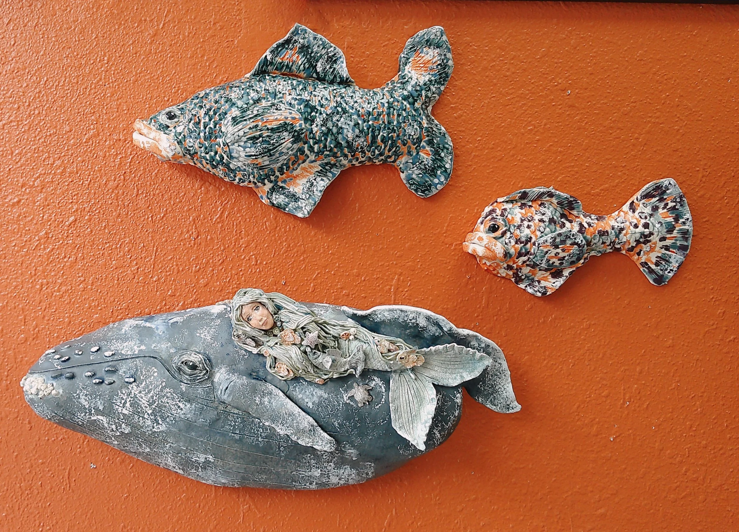 Decorative sea-themed figurines including two fish, a whale with a mermaid figure on its back, and a smaller fish, all on an orange surface.