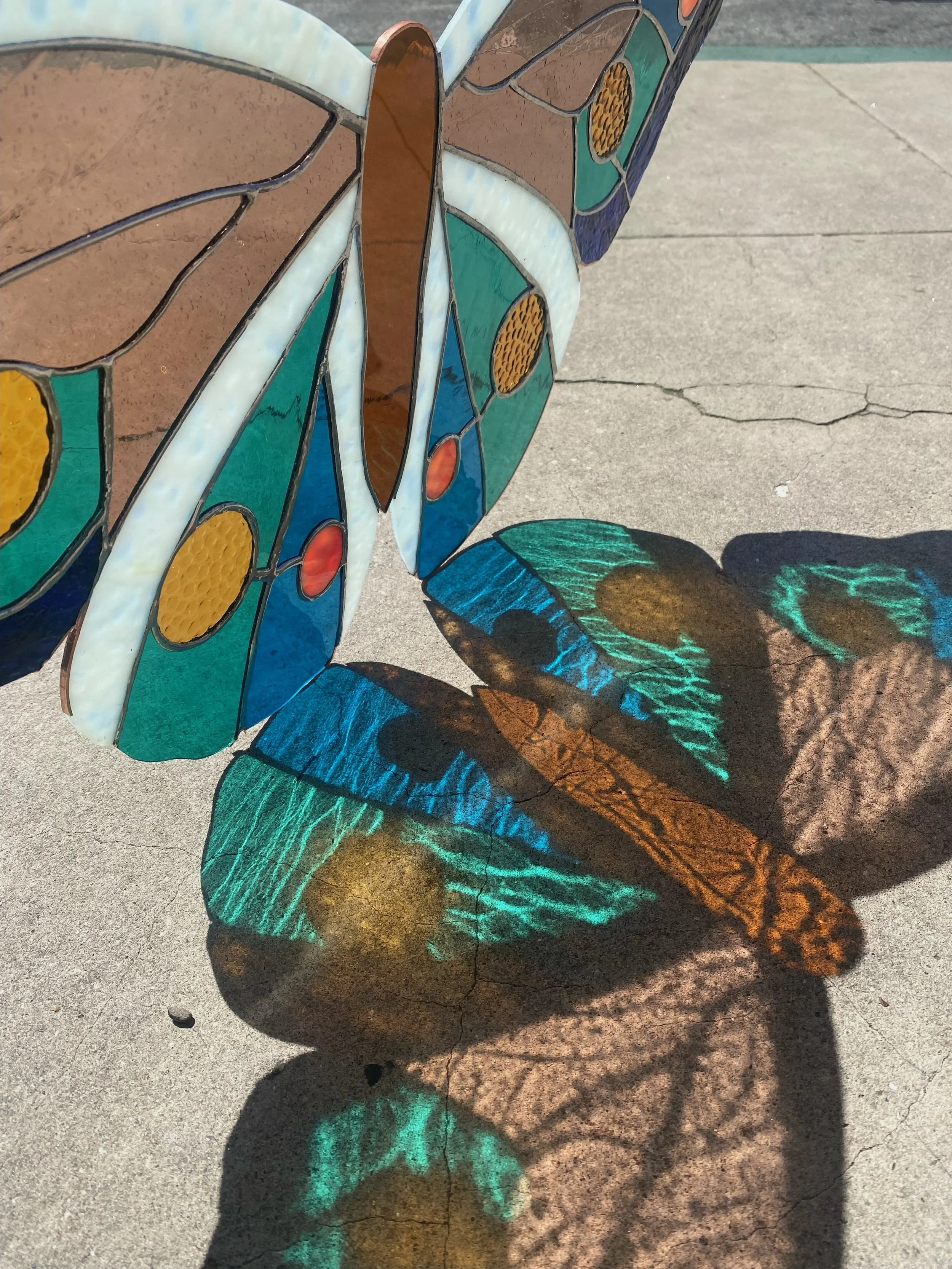 Colorful stained glass butterfly sculpture casting colorful shadows on concrete ground.