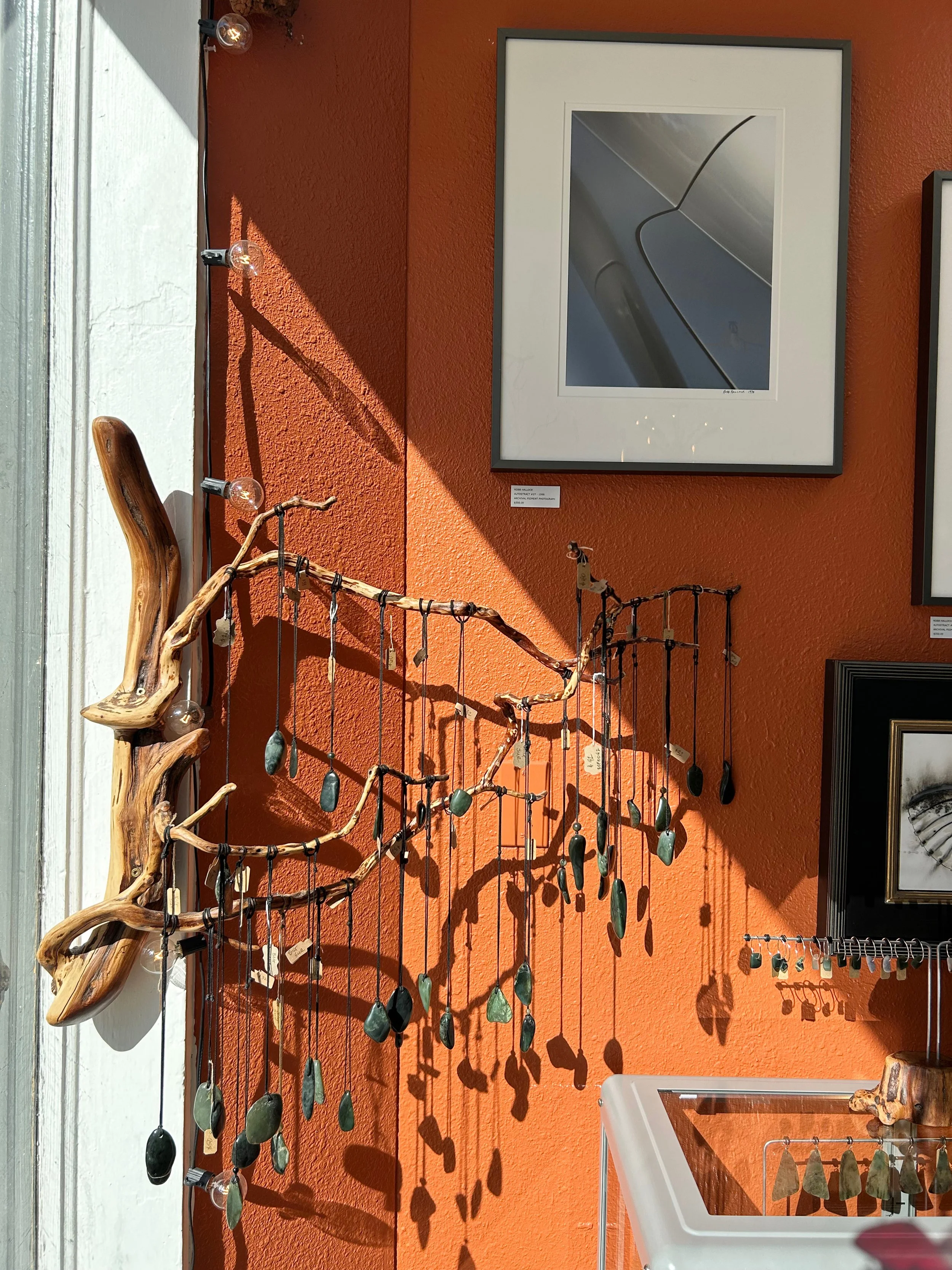 Decorative woodworking and artwork in a gallery display. A piece made of intertwined driftwood with hanging stones, set against an orange wall. Framed modern abstract art on the wall.