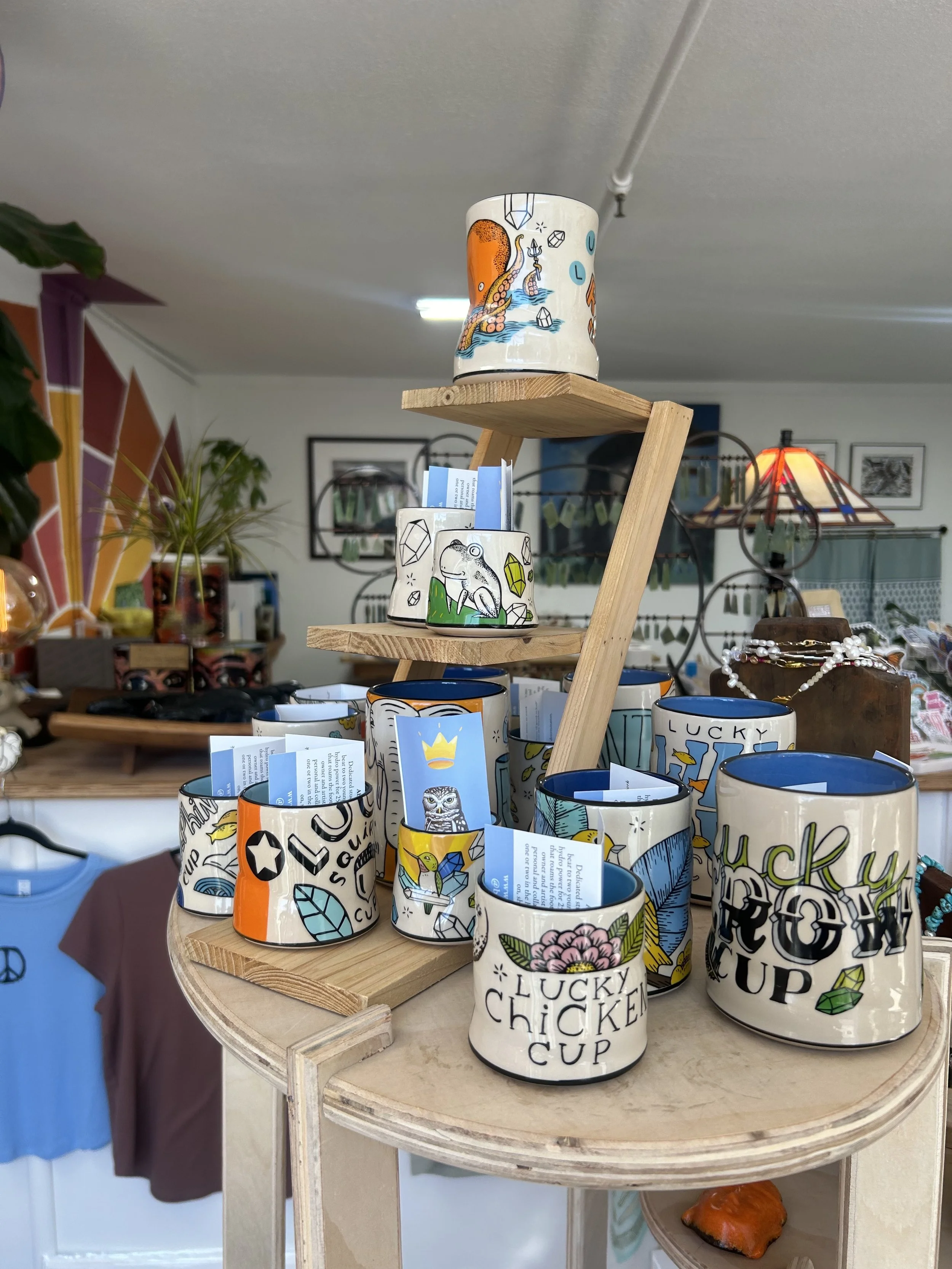 Colorful ceramic mugs with decorative designs and text, arranged on a wooden display stand in a retail store.