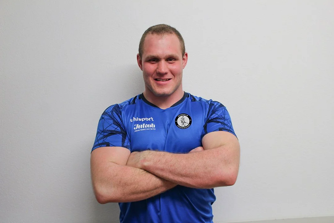 Junger Mann in blauer Sporttrikota, die Arme verschränkt, vor einer weißen Wand, lächelnd.