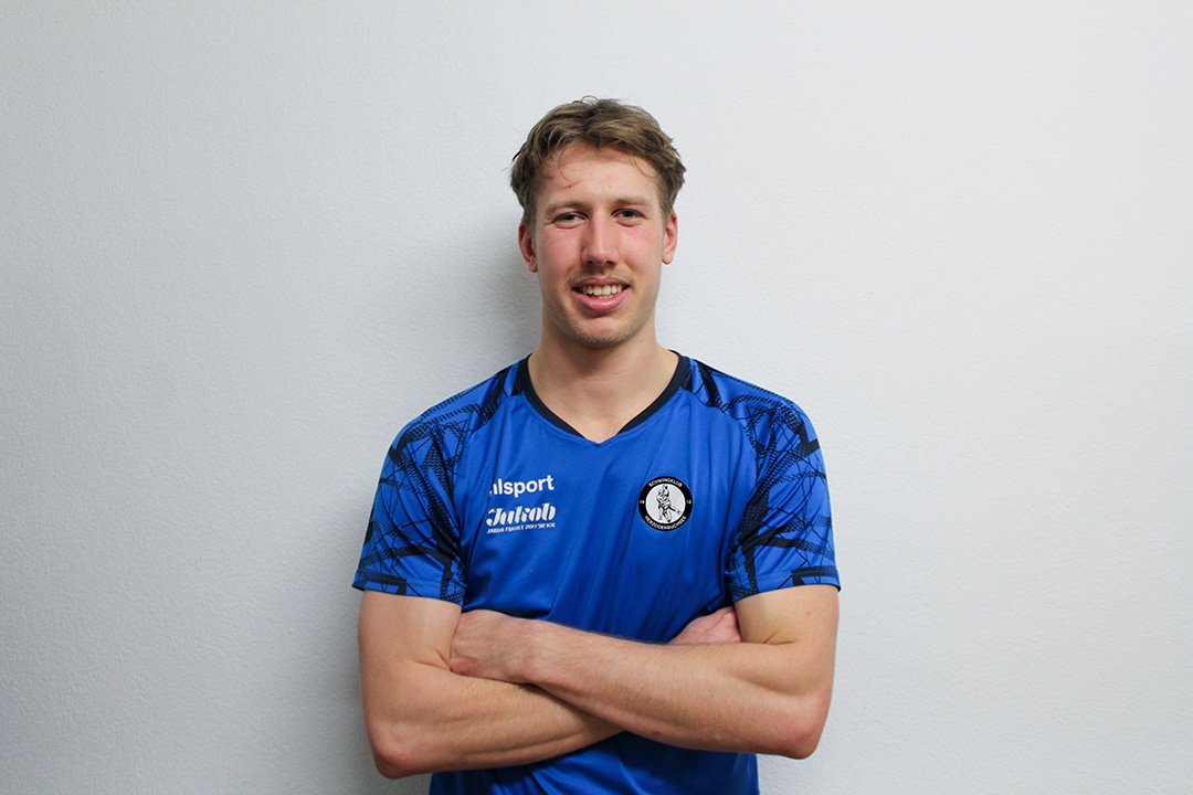 Junger Mann in blauer Sporttrikots mit verschränkten Armen vor weißer Wand.