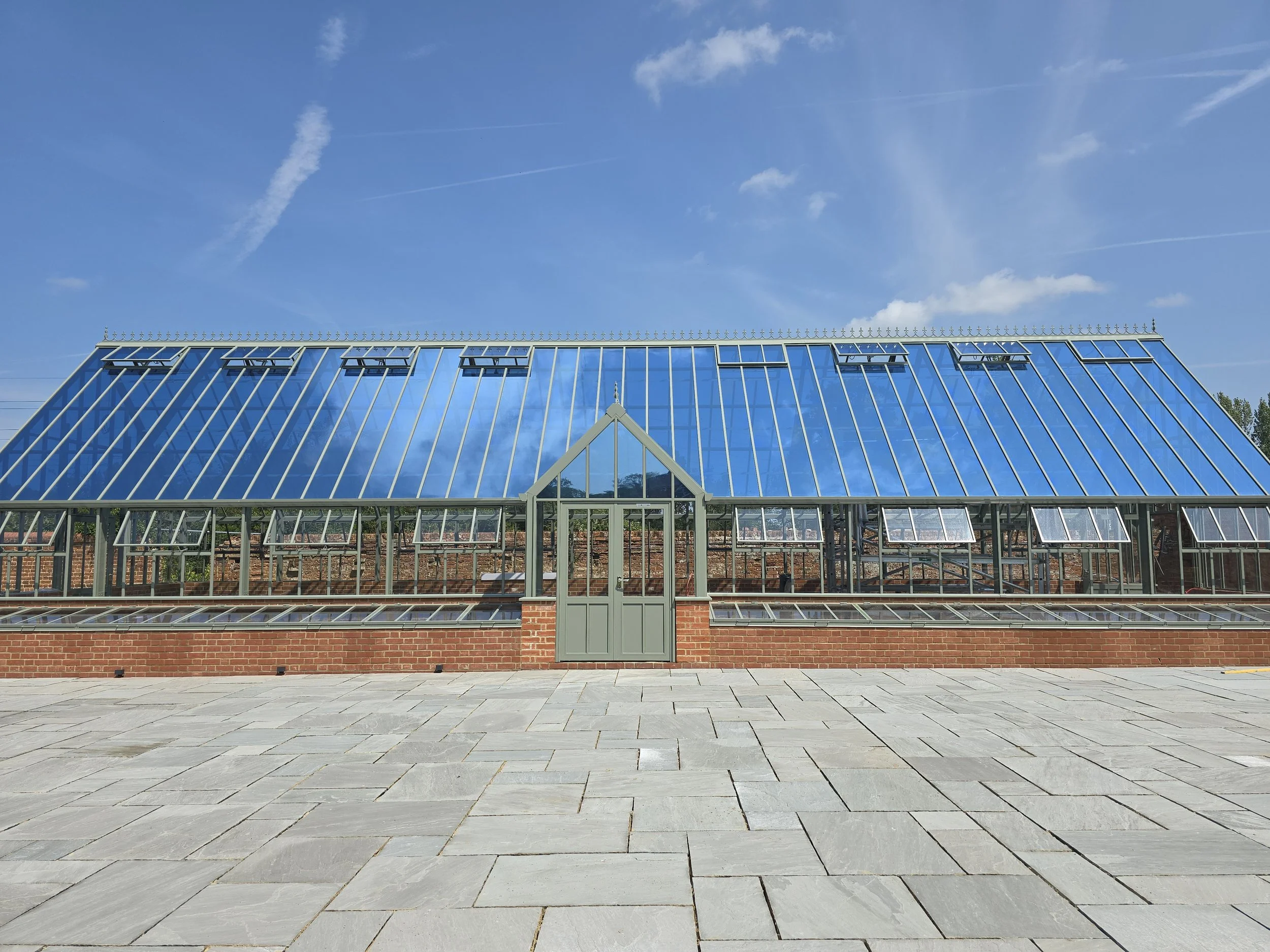 A large greenhouse with a glass roof and brick base, set against a blue sky with wispy clouds.