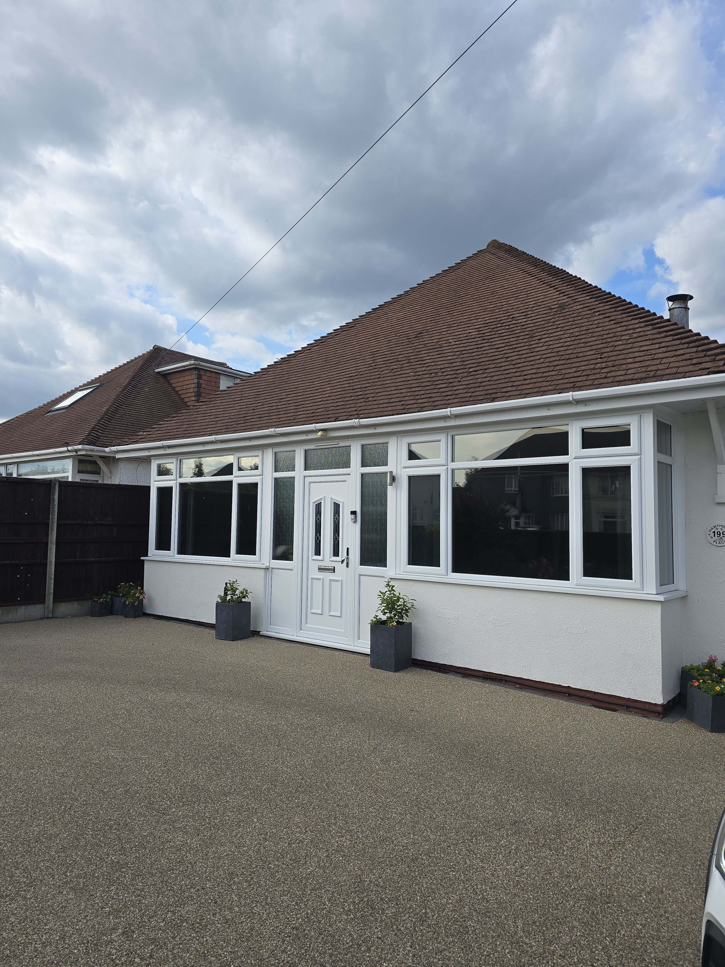 White house with a bay window, and a brown tiled roof, and a closed front door, with potted plants outside, against a cloudy sky.
