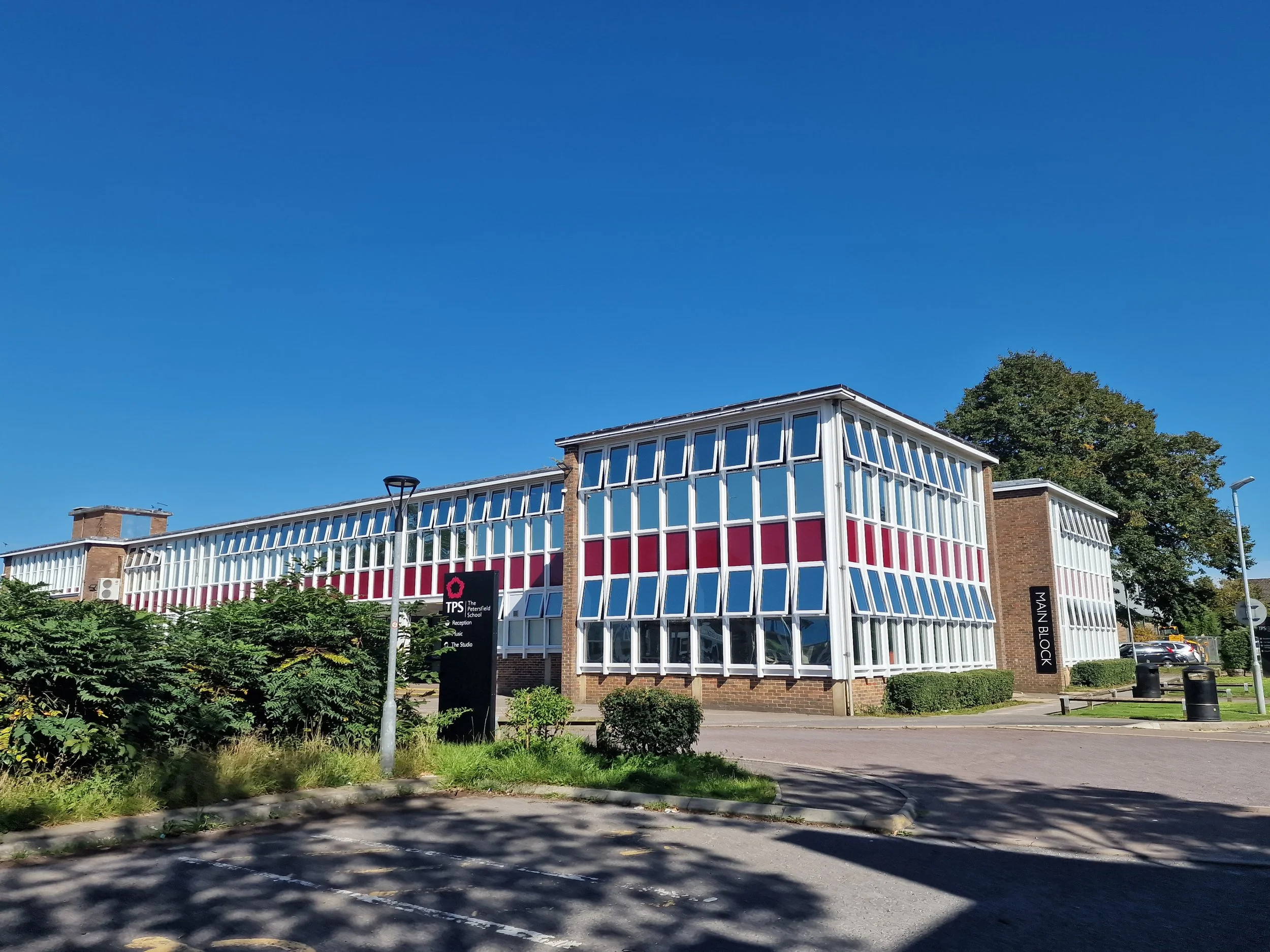 Modern school building with white-framed windows and red accents under a clear blue sky, surrounded by greenery and parking area