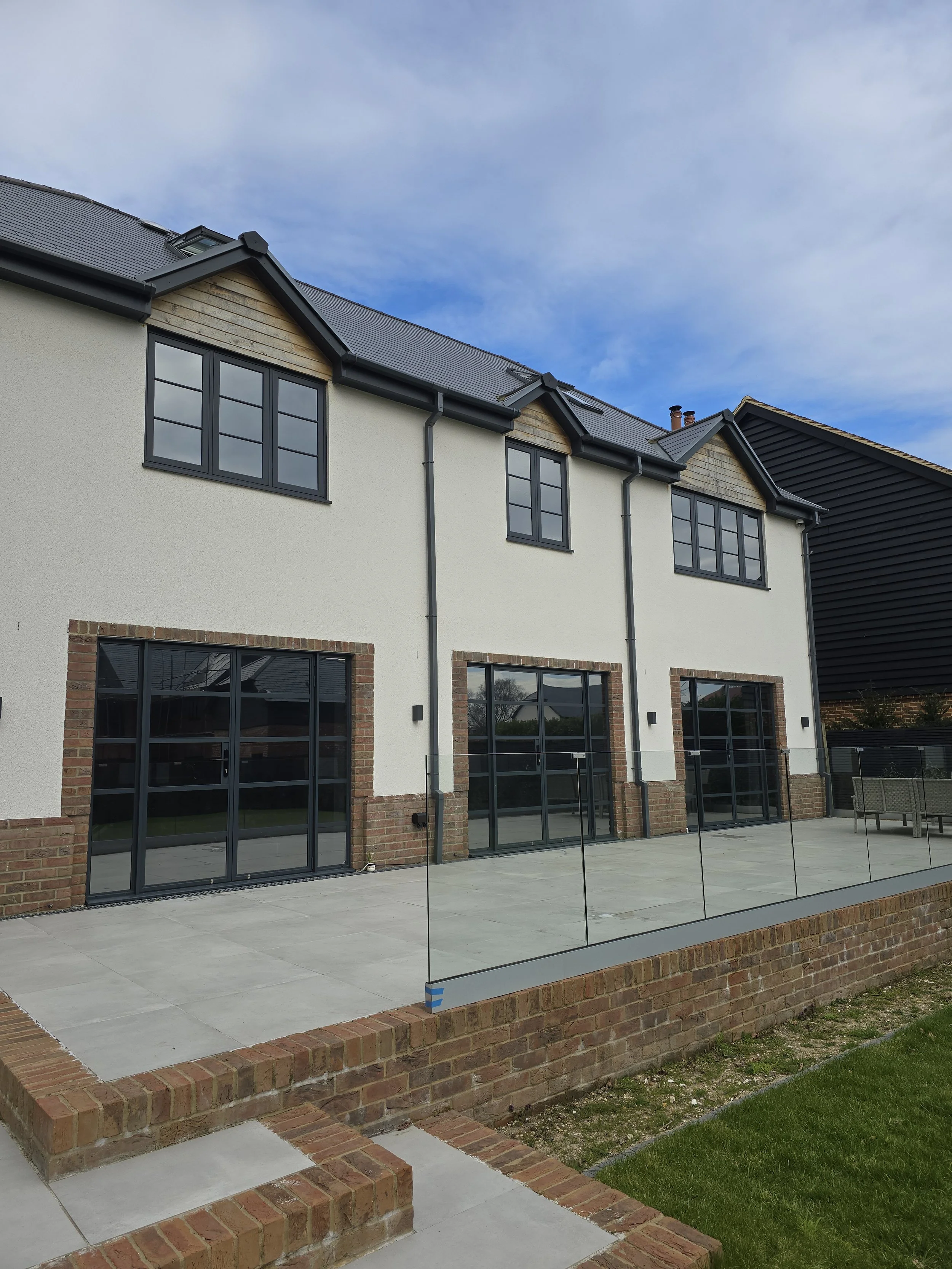 Newly constructed modern house with large glass sliding doors and windows, brick accents, a concrete patio, a brick foundation, and a glass safety fence, against a background of blue sky and neighboring dark-colored building.