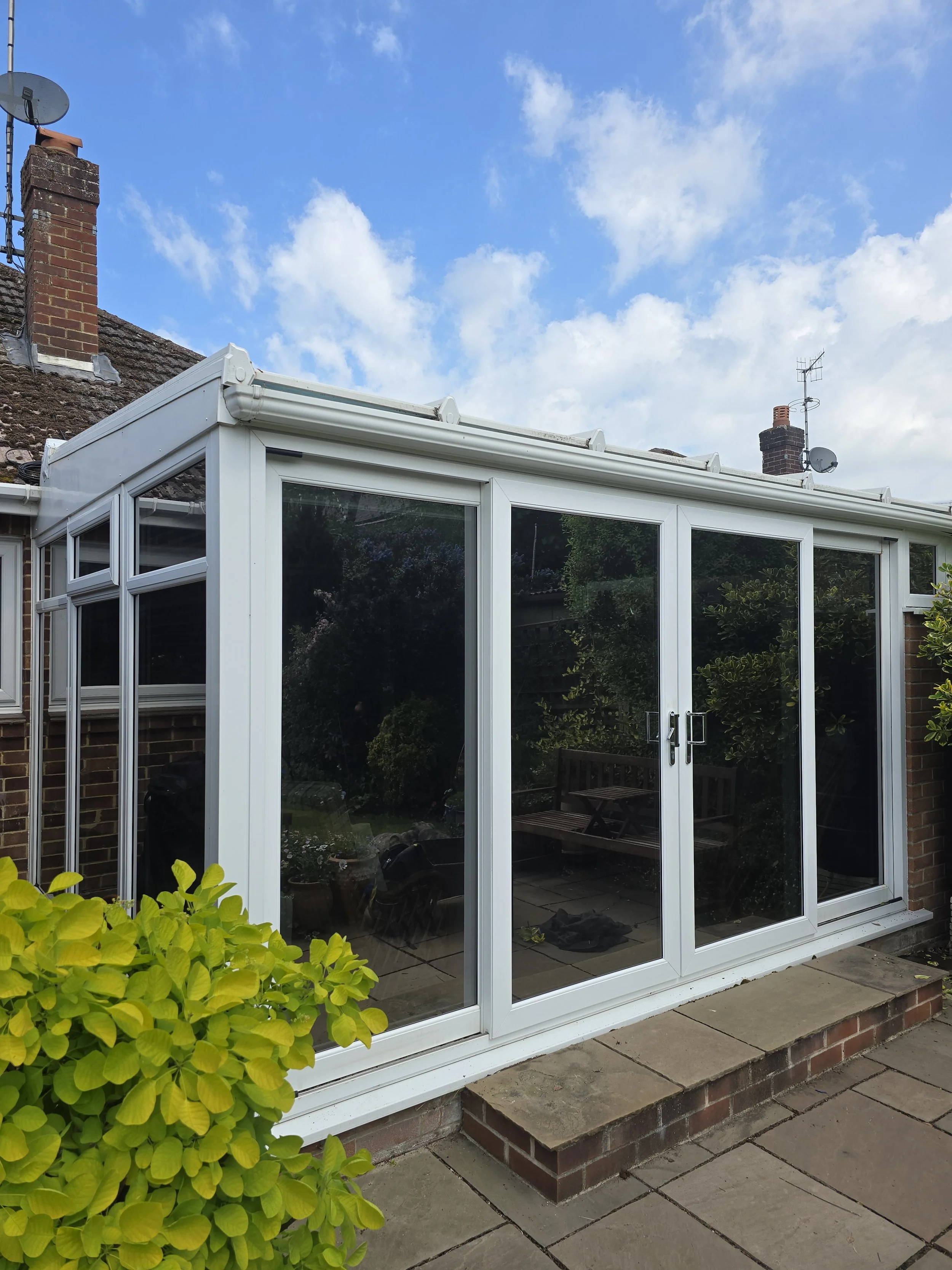 A view of a glass conservatory attached to a brick house, with patio paving stones and a green bush in the foreground, under a partly cloudy blue sky.