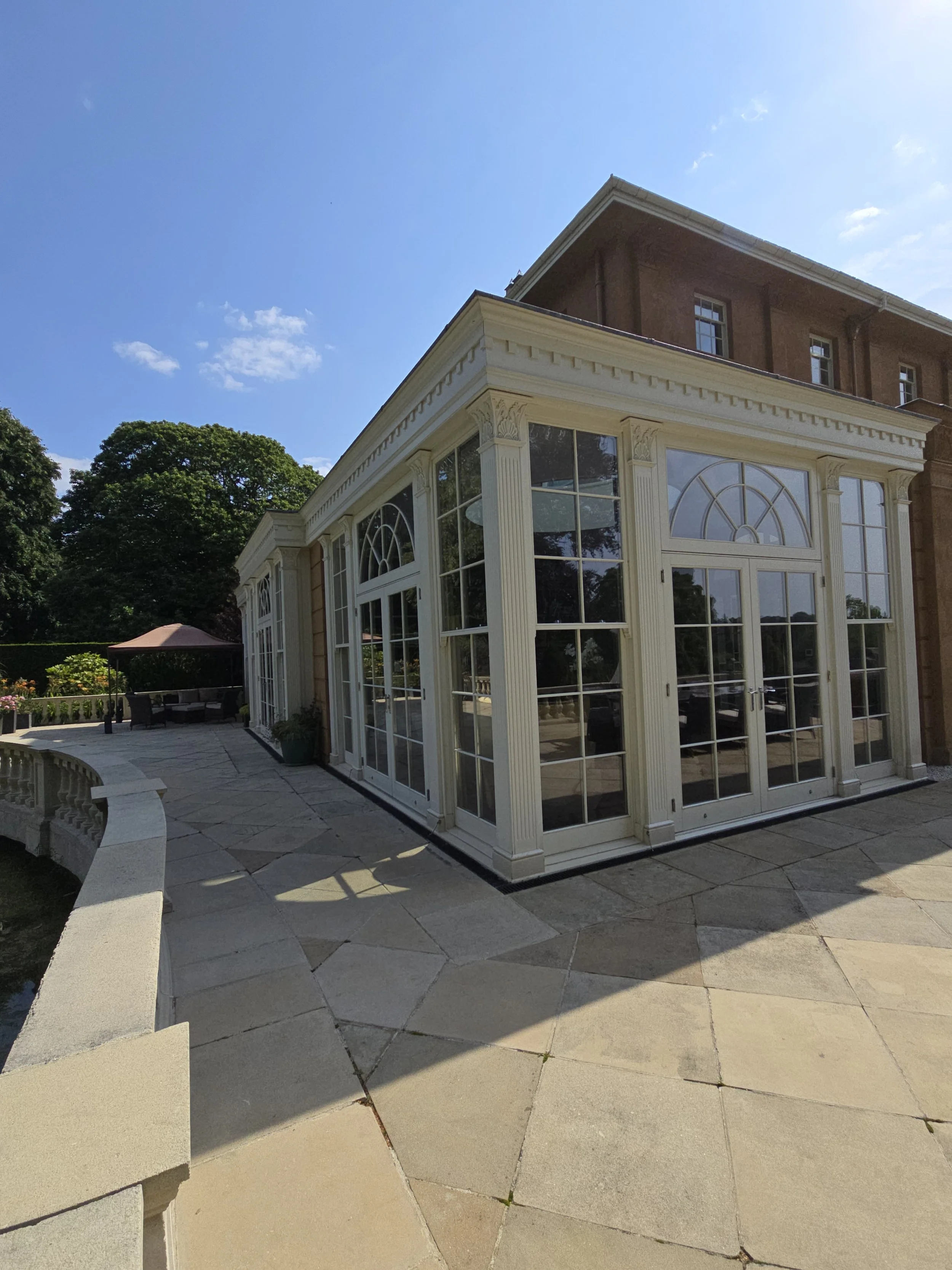 A bright, clear day with a blue sky and a few clouds. A white, ornate porch or conservatory attached to a brick house with large glass windows and doors, surrounded by a landscaped garden with trees and shrubs.