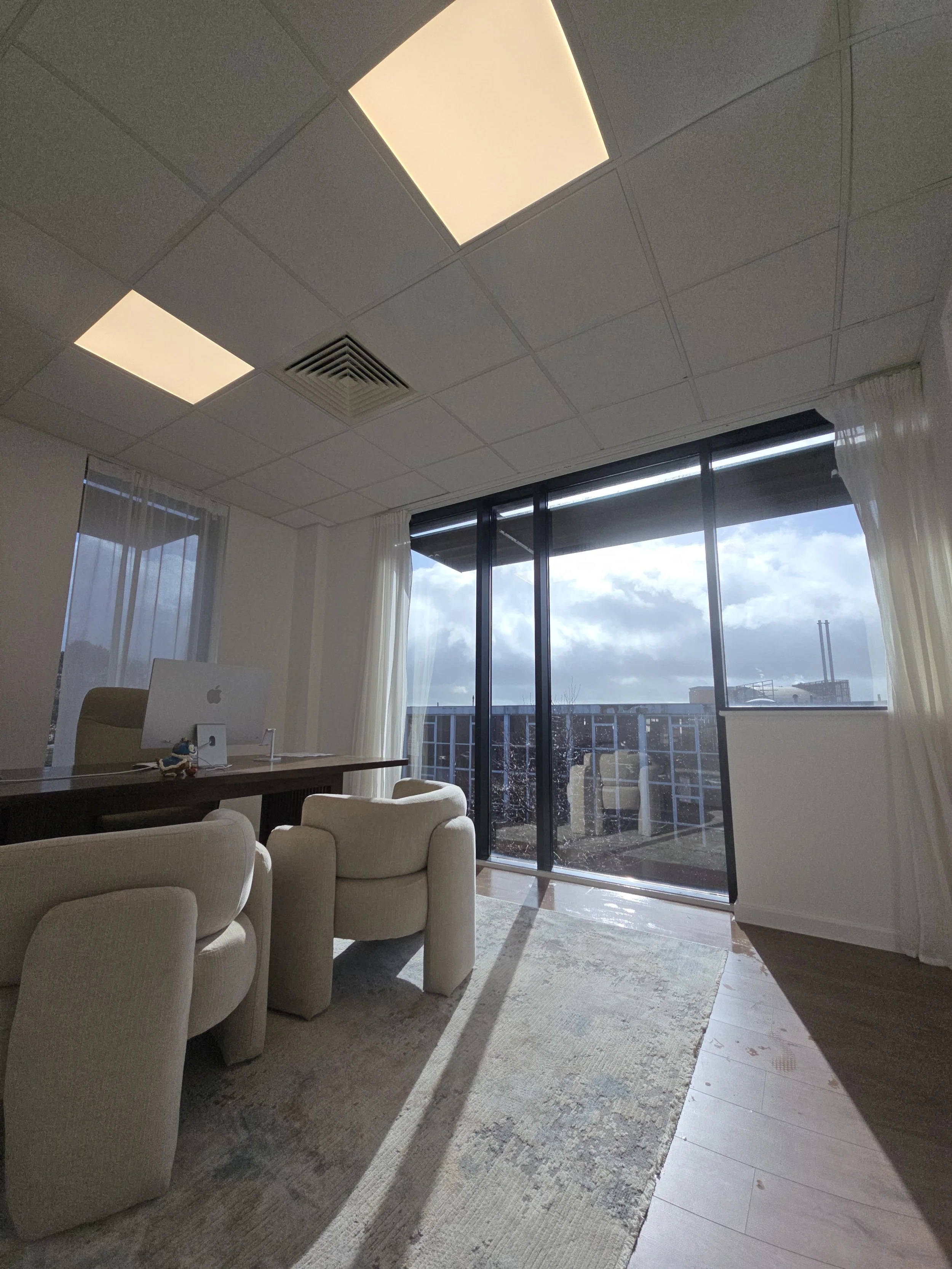 Office space with a desk, chair, and computer near large windows, with sunlight casting shadows on the floor and a cloudy sky outside.