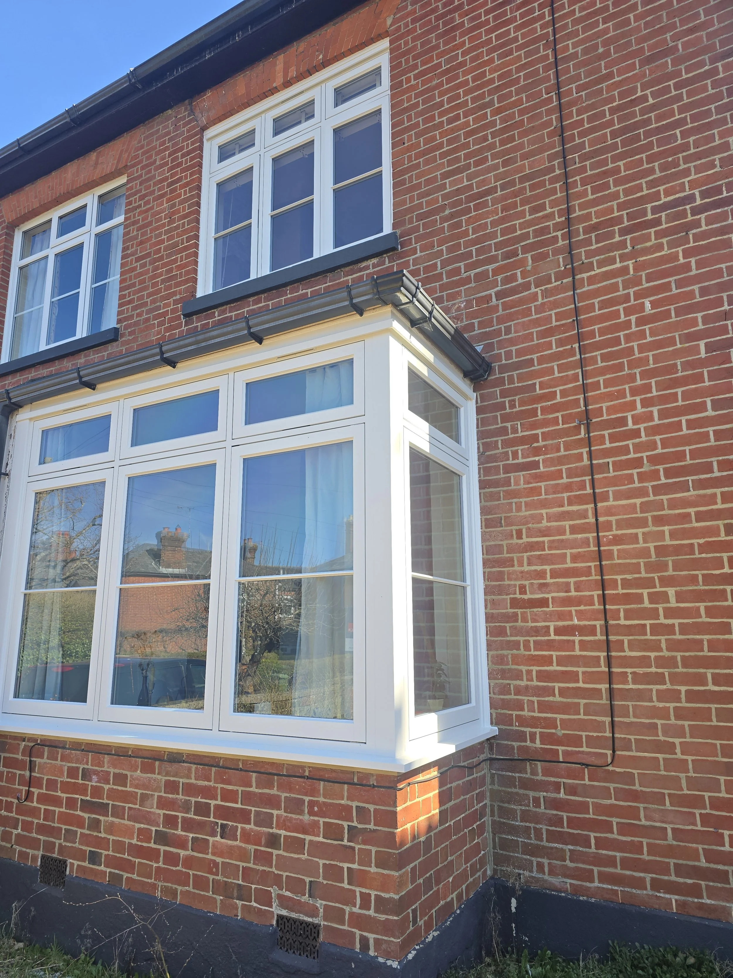 A red brick house with a white bay window, a large front window, and smaller upper windows, with a black roof gutter and a black downspout on the side.