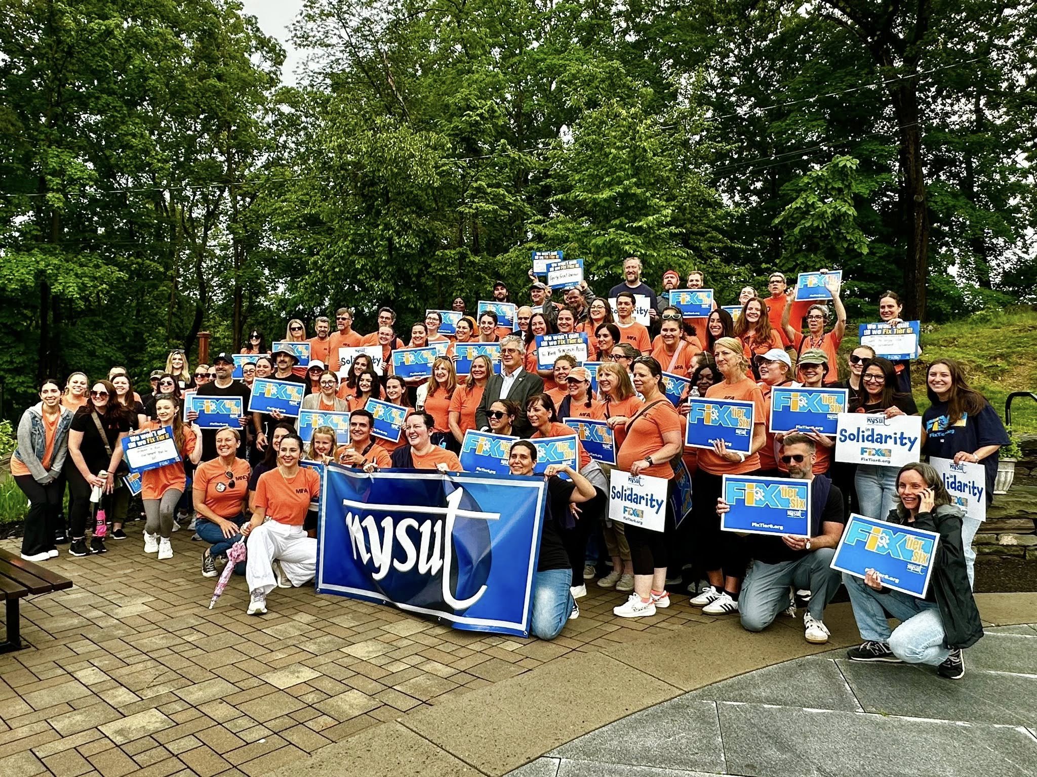 Group photo of diverse people holding signs with the words "Fix the Six," some signs say "Solidarity," outdoors on a paved area with trees in the background.