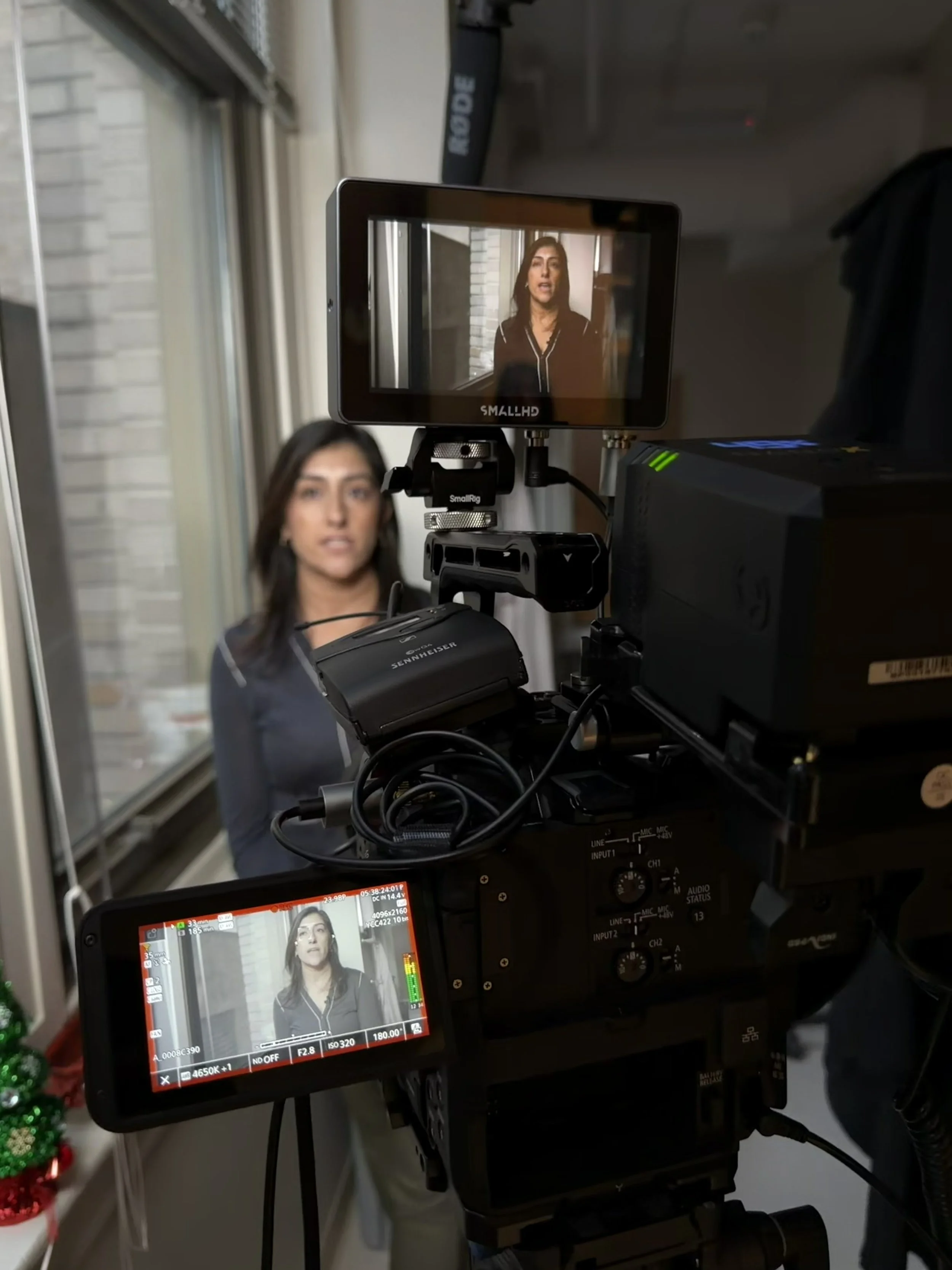 A woman on a film set stands in front of a window, being filmed with a professional camera and monitor showing her in frame.