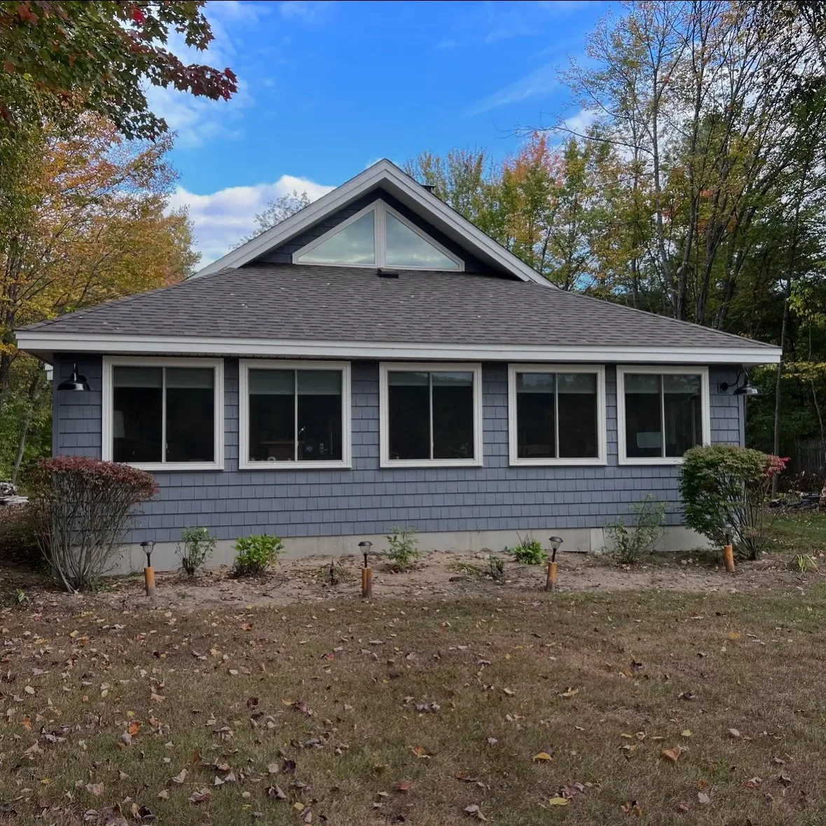 The image shows a house with gray siding and multiple large windows, surrounded by trees with autumn foliage and a yard with small plants and landscape lights.