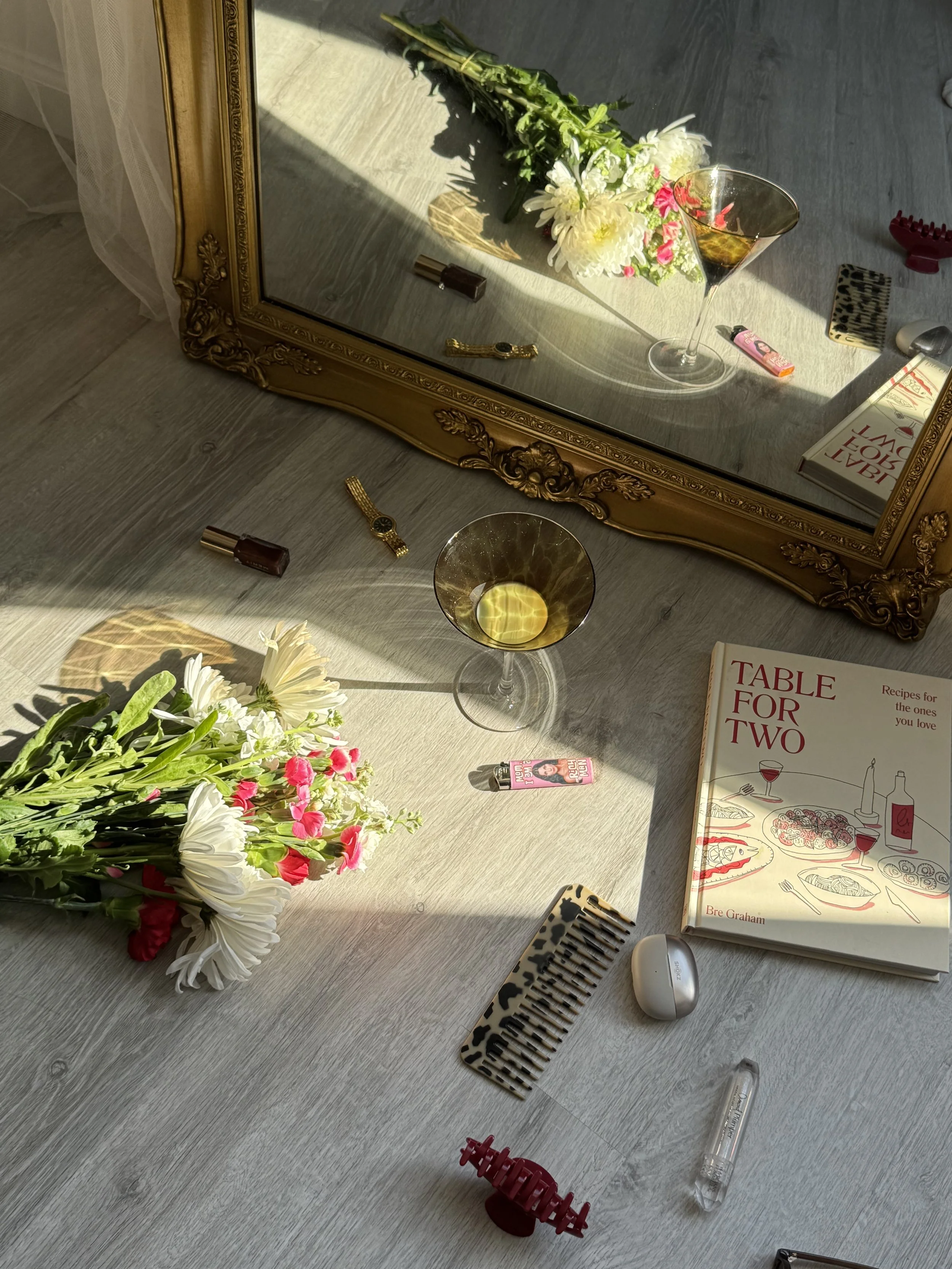 A wooden table with a bouquet of white and pink flowers, a mirror reflecting part of the table, a glass of white wine, a lipstick, a hairbrush, a white wireless earbud case, a glass tube, a book titled "Table for Two," and a leopard print phone case. The table is bathed in sunlight, creating shadows.