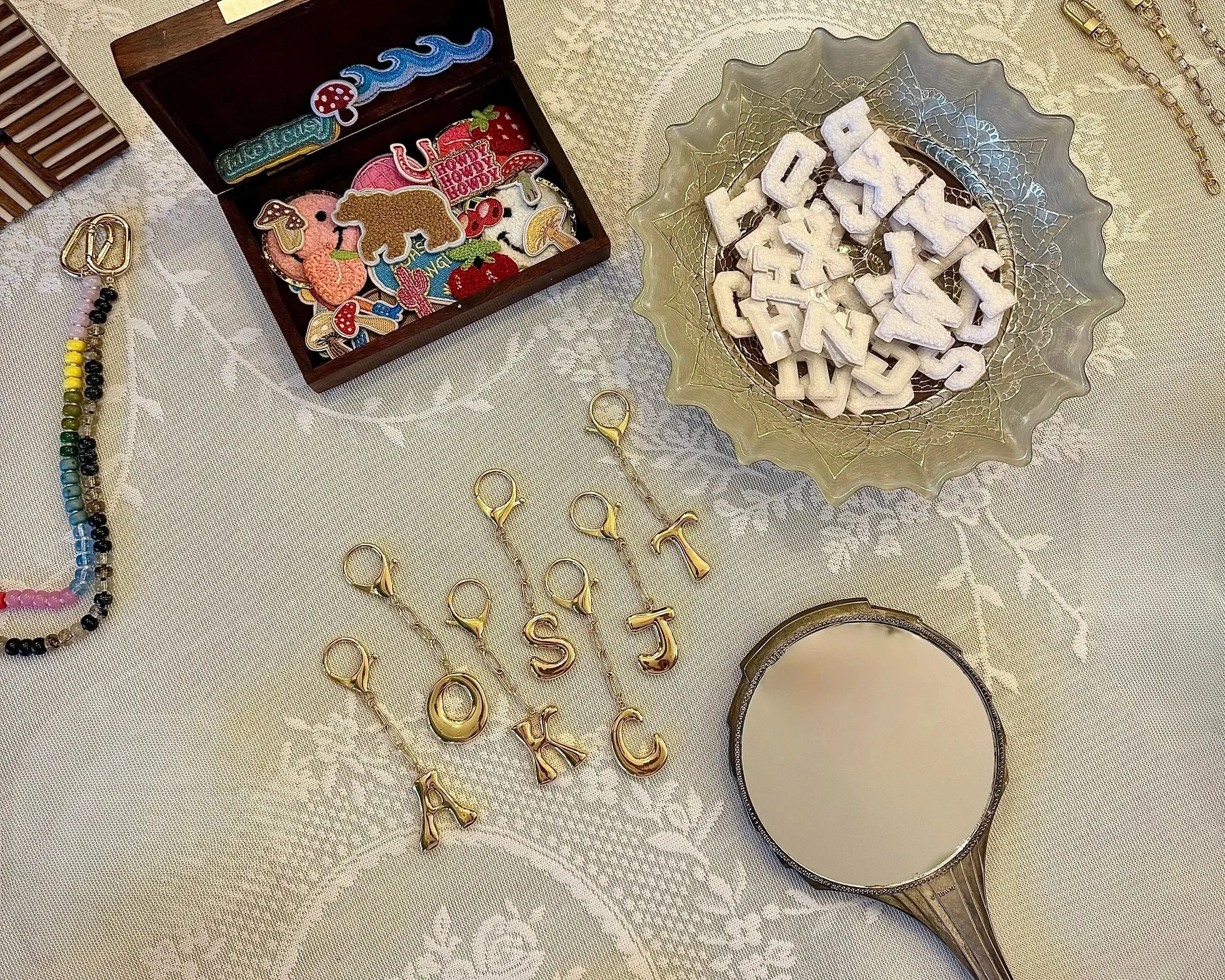 Decorative jewelry and accessories including a jewelry box with patches and embroidered designs, a glass bowl with white foam letters, gold earrings, and a mirror on a lace tablecloth.