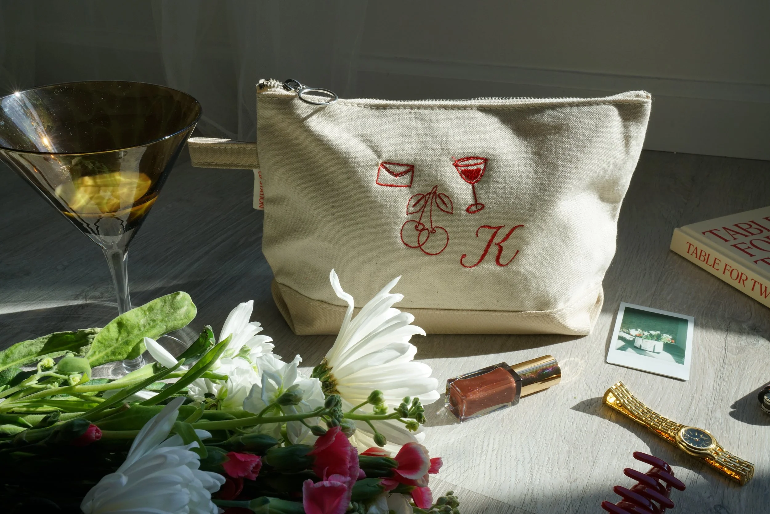 A beige cloth zippered pouch with red embroidered illustrations of mail, a wine glass, cherries, and the letter 'K', surrounded by a bouquet of white and pink flowers, a gold wristwatch, lipstick, and scattered makeup items on a gray surface.