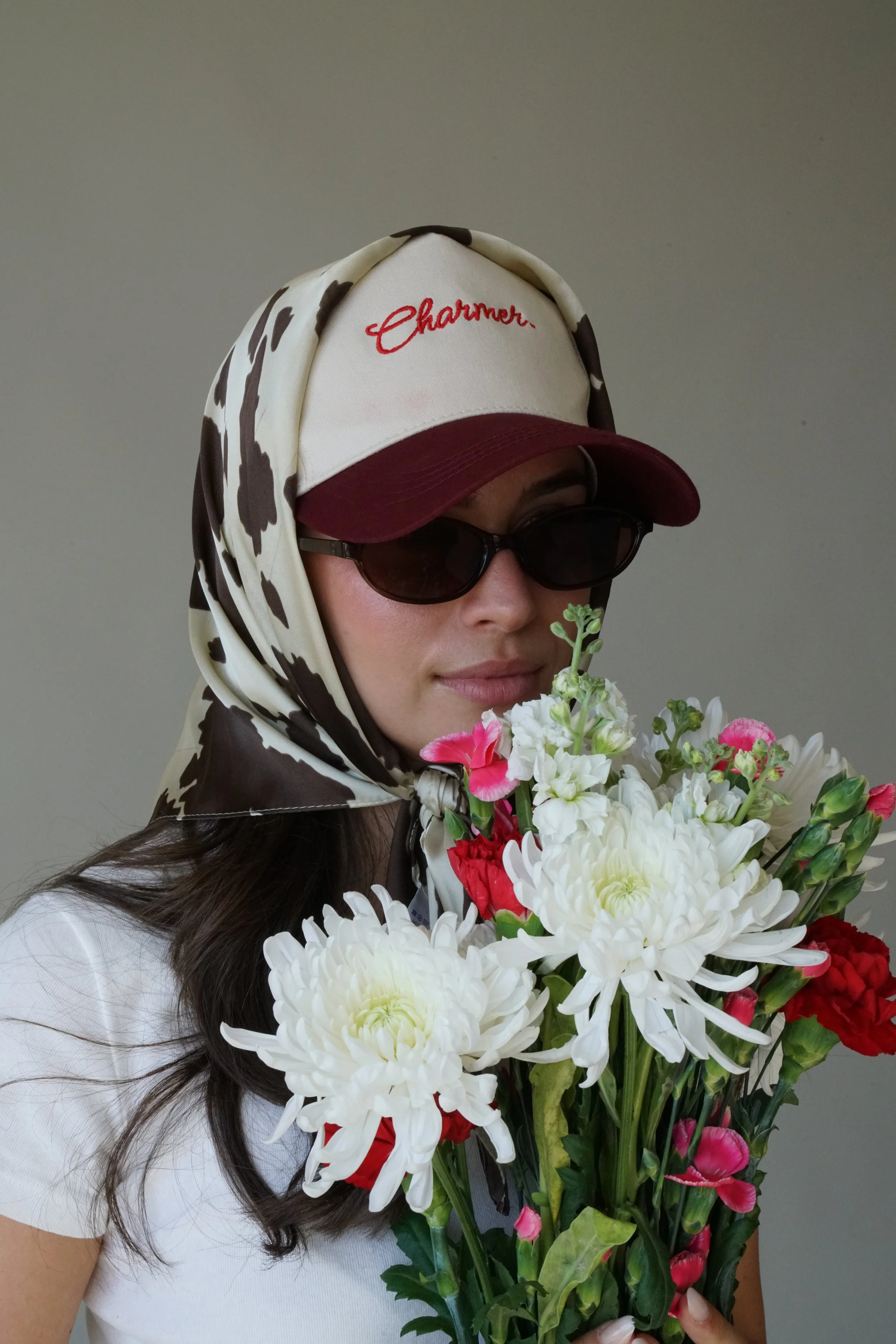 A woman wearing dark sunglasses, a red and white baseball cap with the word "Charmer" embroidered on it, a patterned scarf over her head, and a white shirt, holds a bouquet of mixed flowers including white chrysanthemums, pink, and red blossoms.