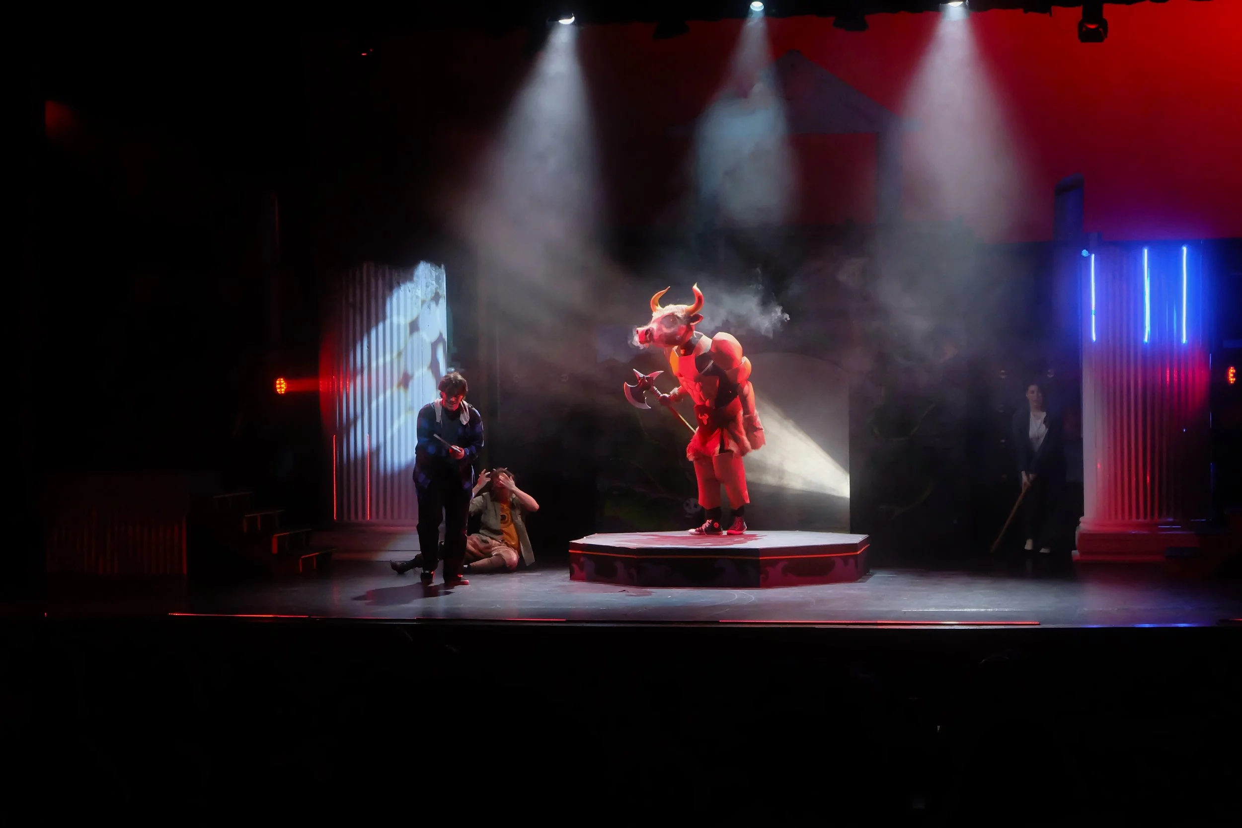 A stage performance featuring a person in a demon or devil costume with horns, a mask, and an axe prop, standing on a platform, with other performers or crew members seated or standing nearby, illuminated by spotlights and stage lighting.