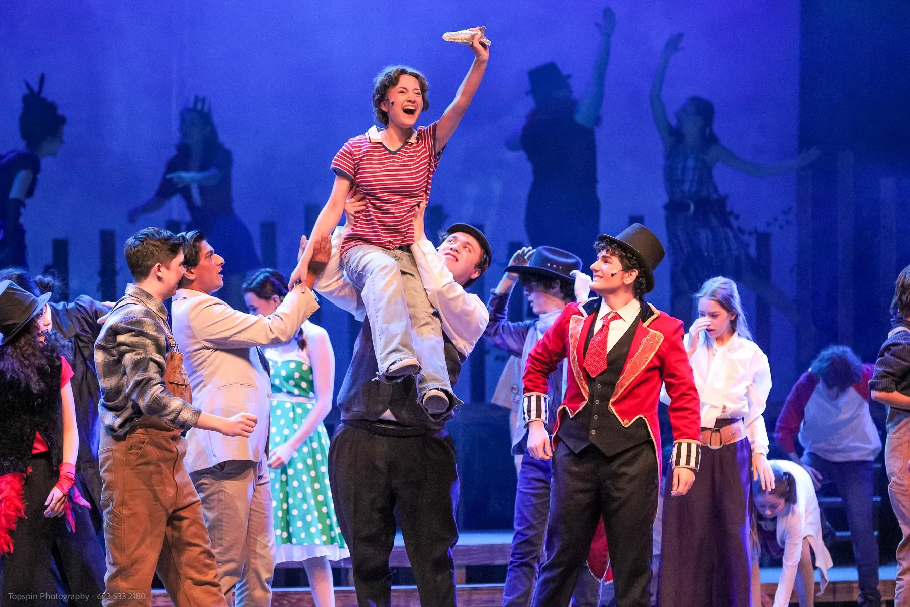 Actors on stage performing a lively musical with a group of children dressed in colorful costumes, with one girl being lifted up and smiling amid the crowd.