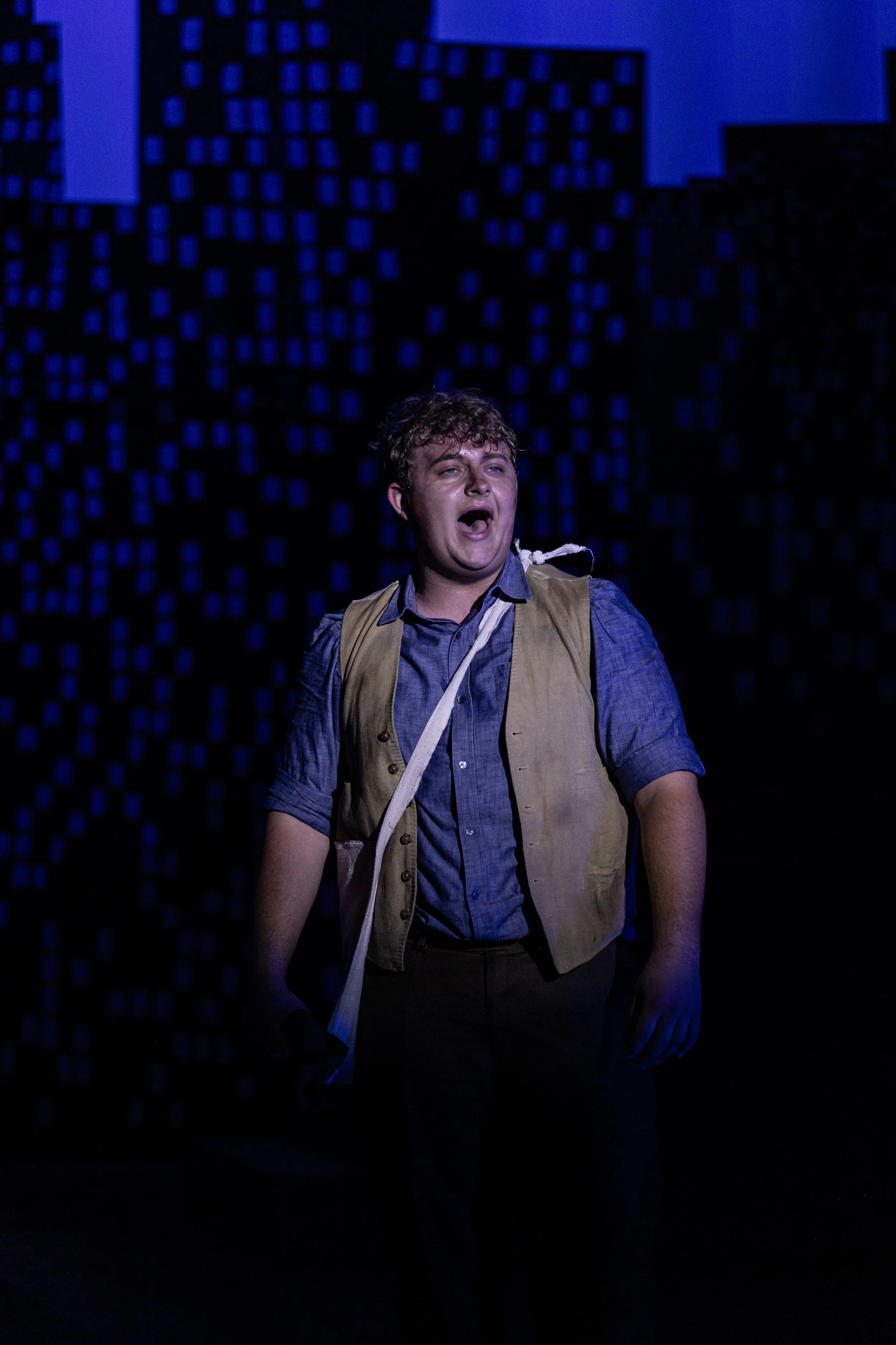 A person with curly hair singing or shouting on a dark stage with a blue-lit digital cityscape background.