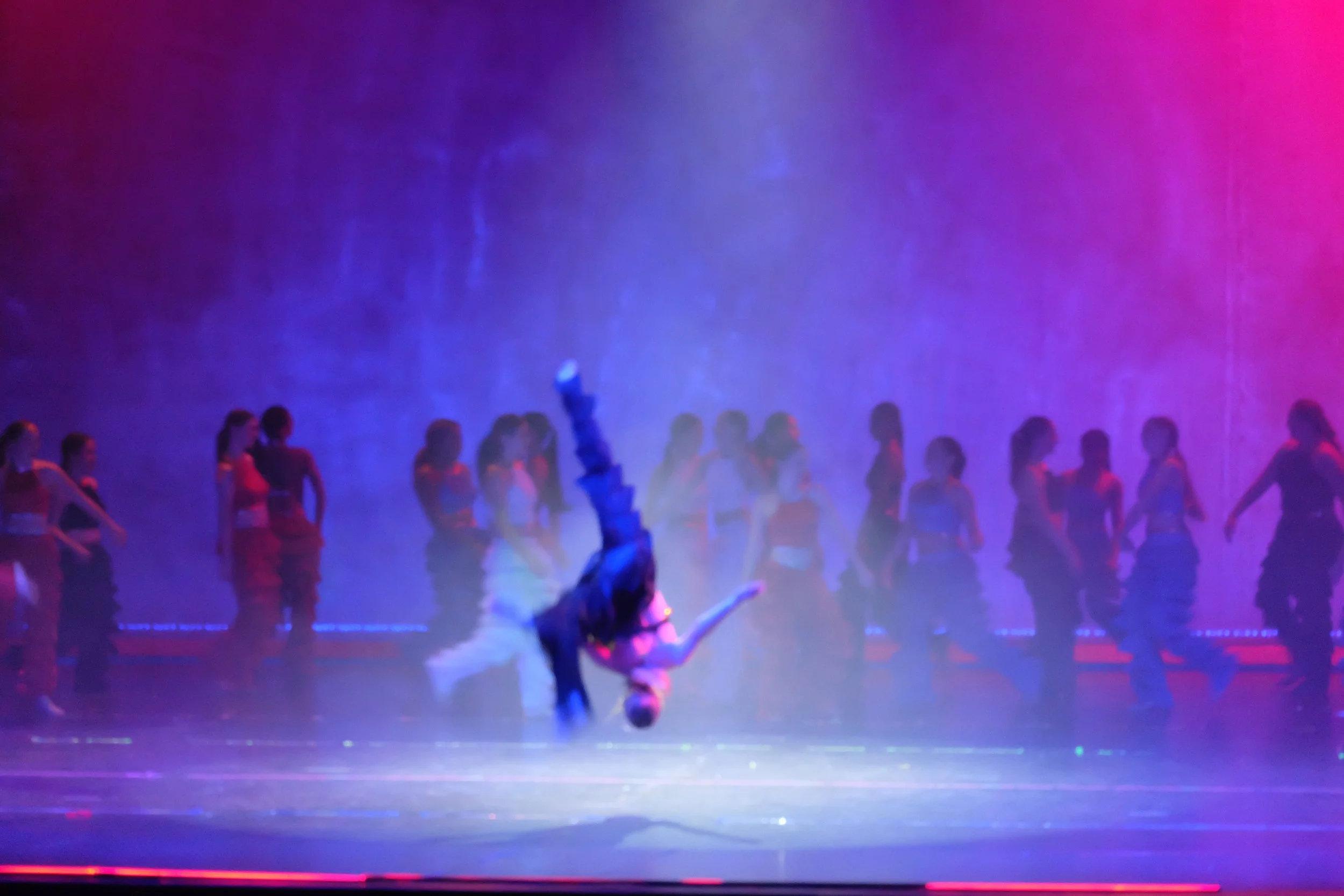 A performer is upside down on their hands in a dance or acrobatic performance on a stage with colorful purple and pink lighting and a group of people dressed in costumes or uniforms in the background.
