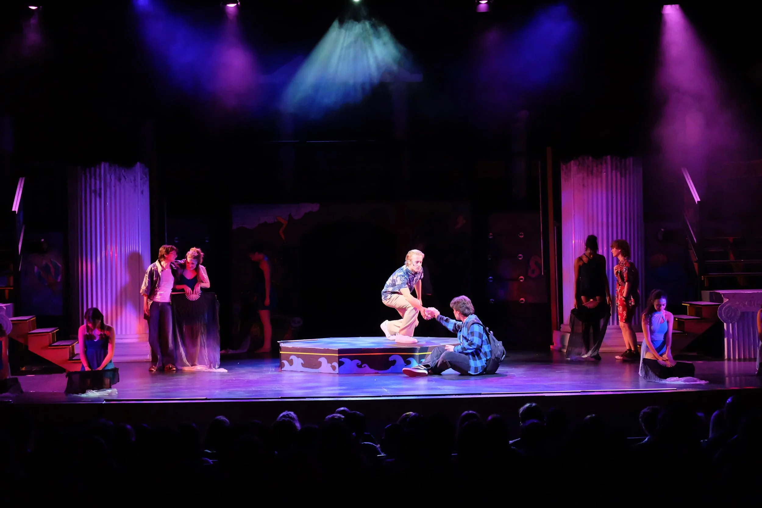 Stage performance with actors and dancers under purple and yellow spotlights.