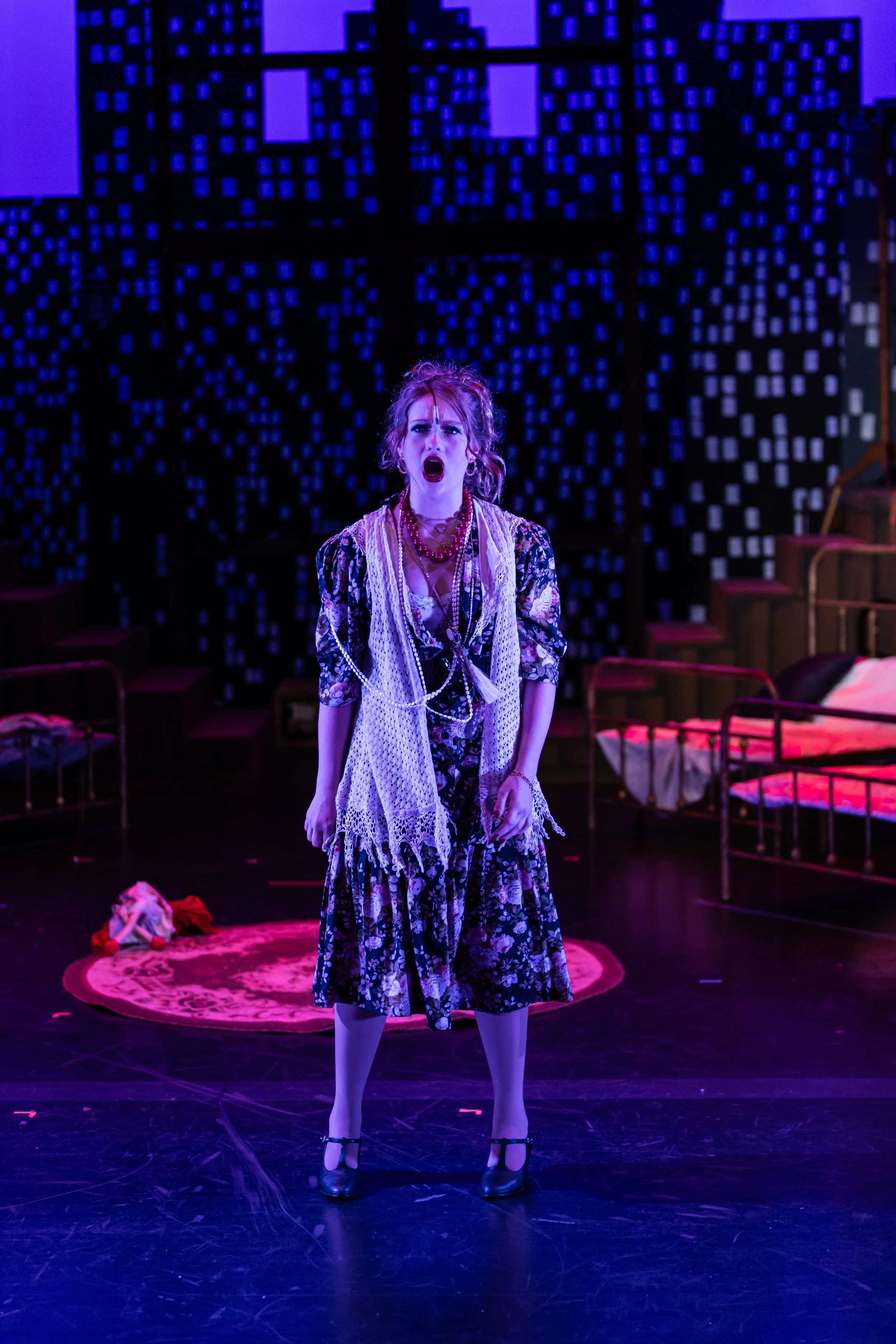 A woman with curly hair singing or shouting on a stage with a blue and black geometric patterned background. She is wearing a floral dress, a white lace vest, red necklaces, and black shoes. There are chairs and a doll on the stage floor.