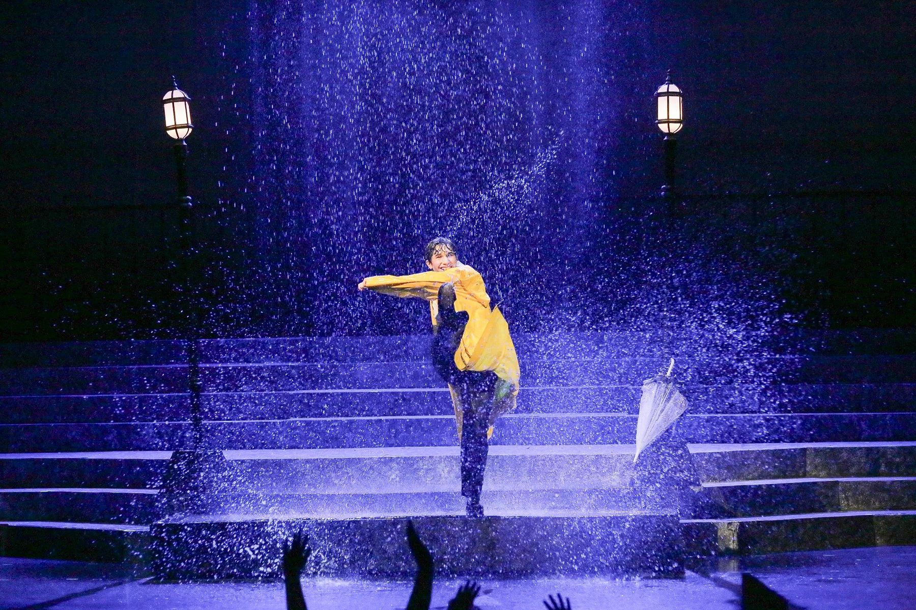 A performer wearing a yellow raincoat dances in the rain while holding an umbrella on stage at night. Stage lights and stairs are visible in the background, with audience hands raised in the foreground.