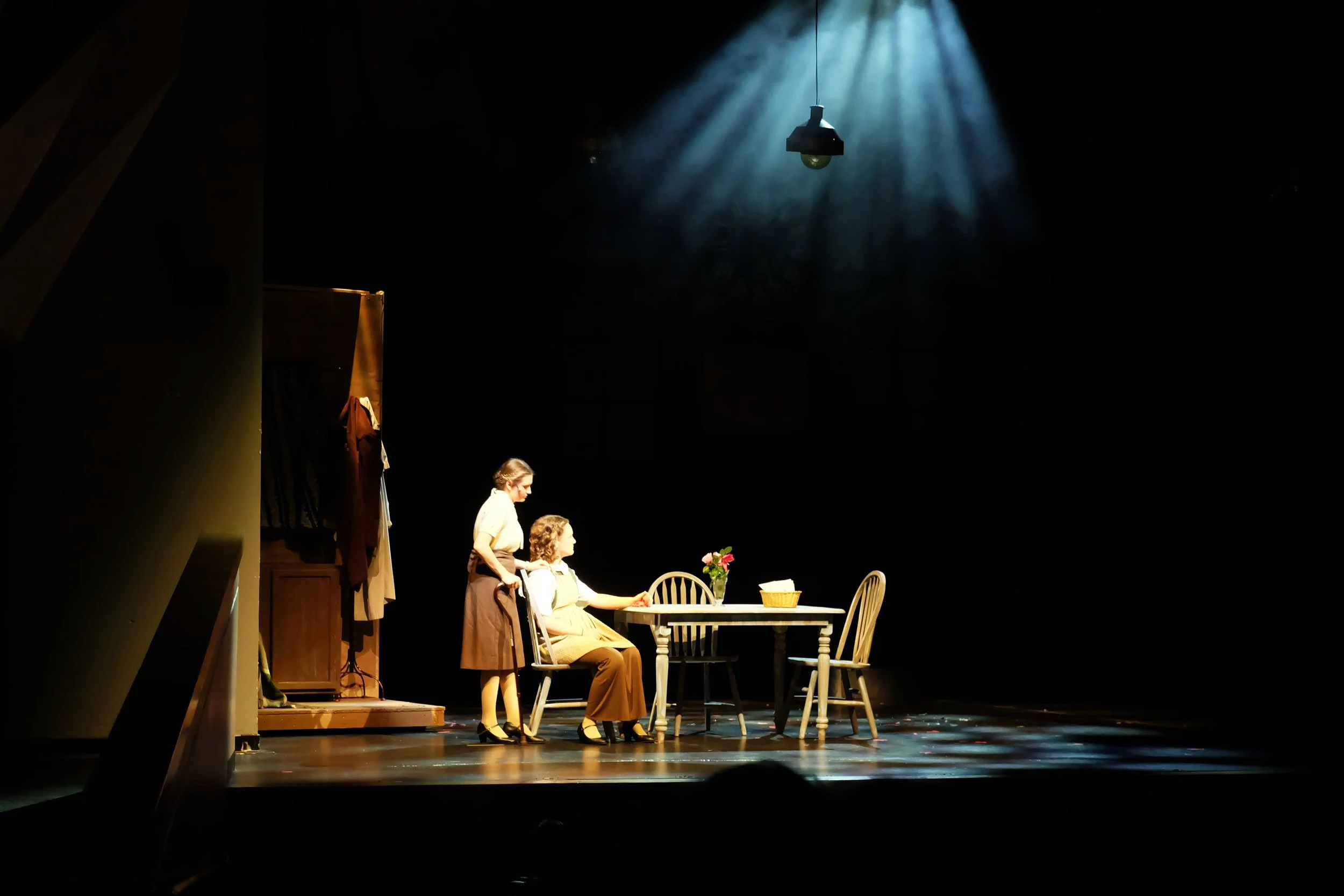 The image depicts a stage scene with a woman sitting at a table with a vase of flowers, and two women standing beside her, all dressed in vintage clothing. The stage is dimly lit, with a single overhead light shining down, creating a dramatic atmosph