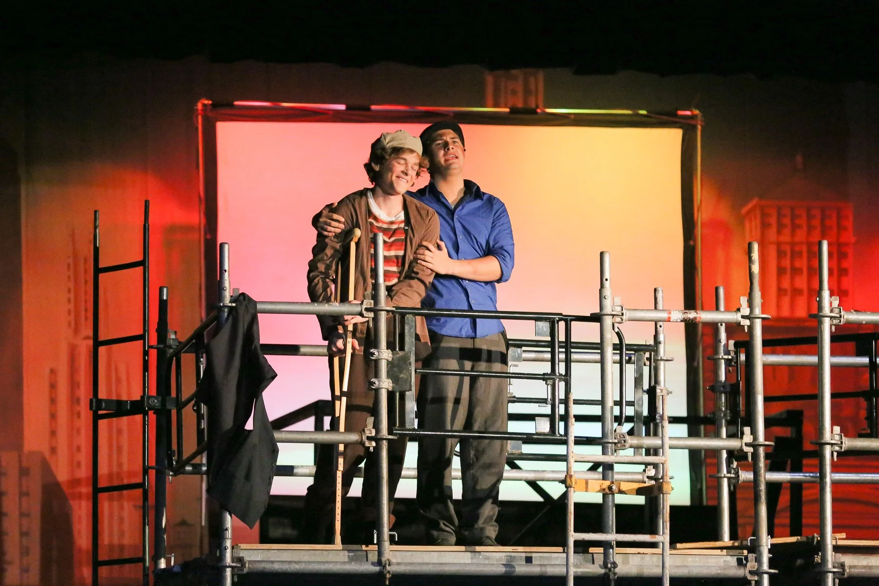 Two actors stand on a stage behind scaffolding, one smiling and the other appearing emotional, with a large screen and cityscape backdrop behind them.