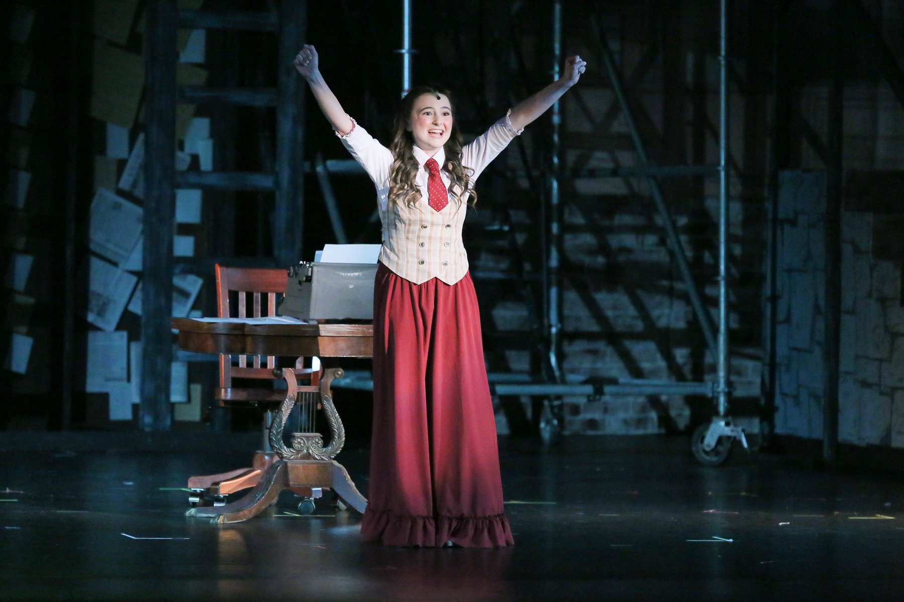 A young woman on stage with arms raised, smiling, dressed in vintage clothing including a white shirt, red tie, beige vest, and long red skirt. There is a desk with papers, a typewriter, and a chair behind her. The stage background is dimly lit with 
