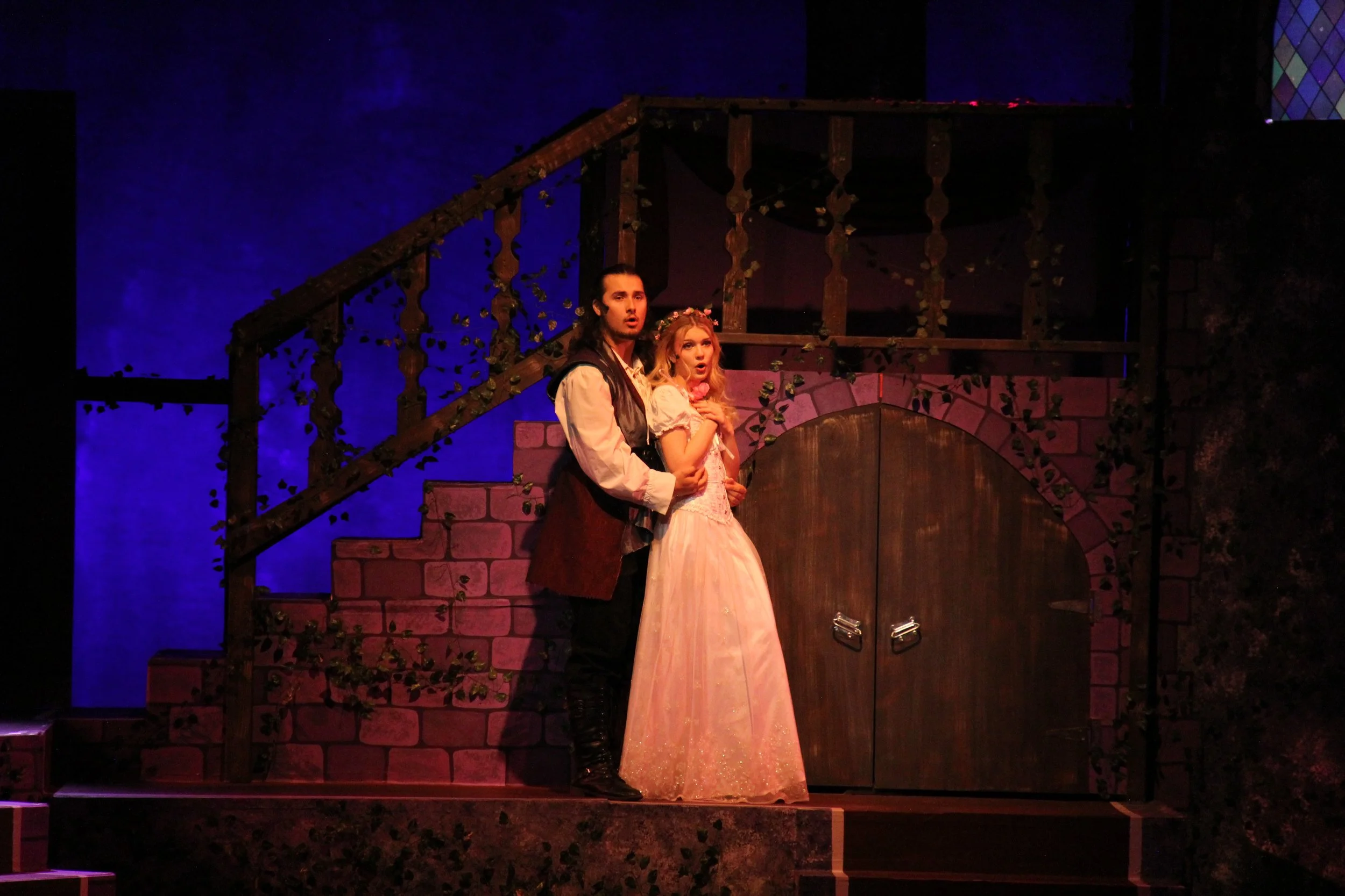 Two actors on stage, a male and female, dressed in medieval style costumes with a castle backdrop, during a theatrical performance.