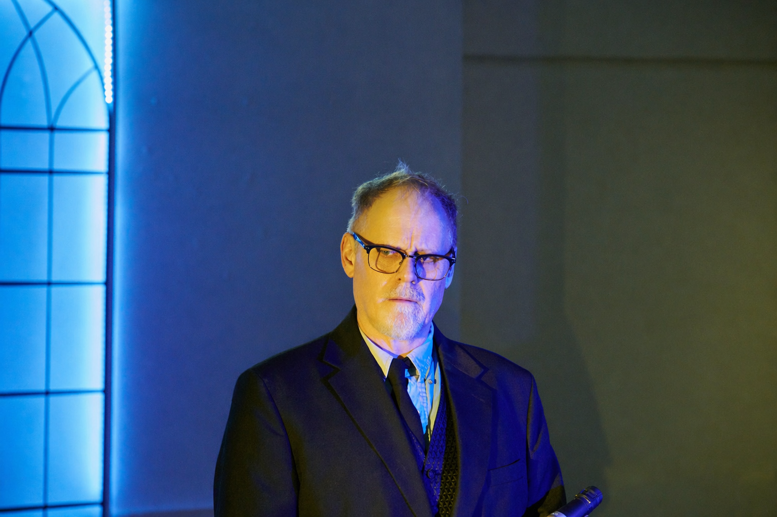 A man in a dark suit and glasses stands in a dimly lit room with blue and yellow lighting, holding a microphone.