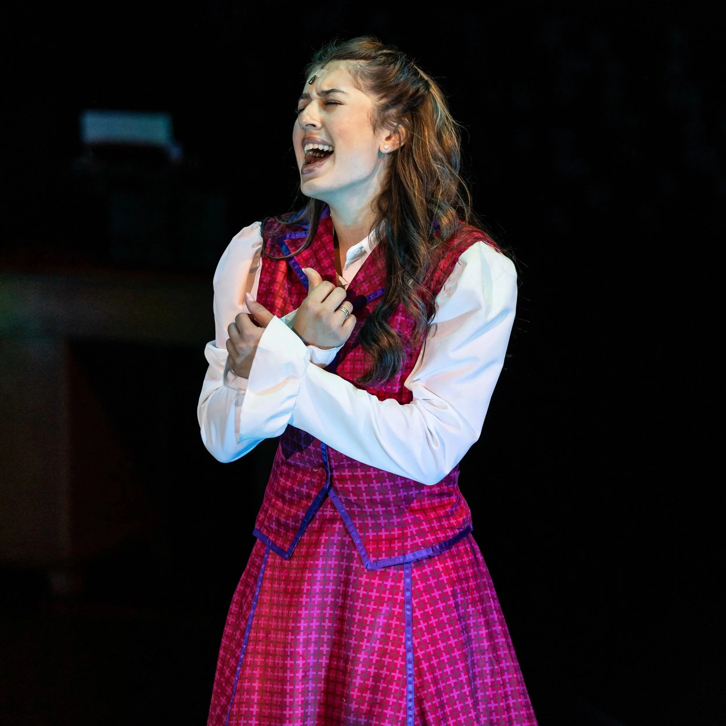 A young woman with long wavy hair wearing a white shirt and a red checkered apron stands against a dark background, singing or speaking with her eyes closed and mouth open, holding her chest with her left hand.