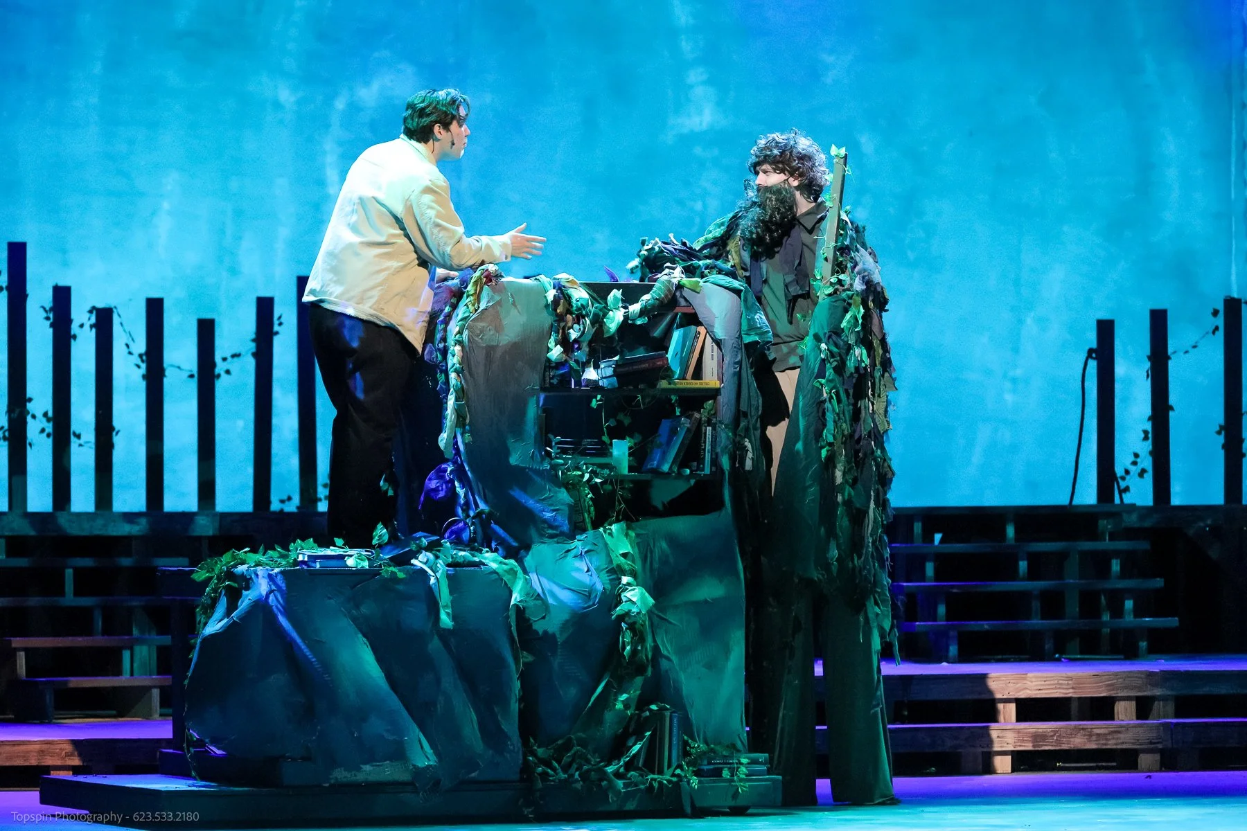 Two actors on stage engaged in a scene, with one standing and gesturing towards the other, who is dressed in a costume resembling a bush or tree, surrounded by props and set pieces that suggest an outdoor or natural setting.