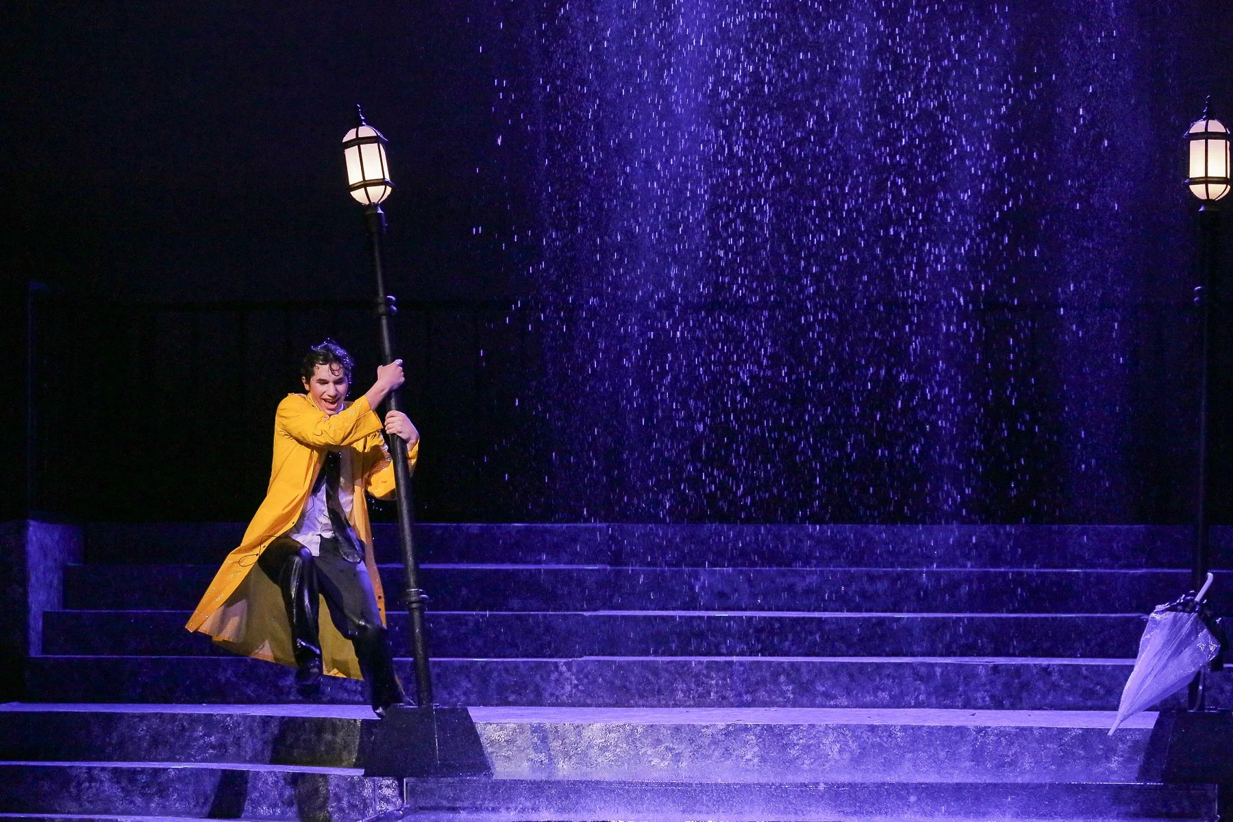 Person dressed in a yellow raincoat holding onto a street lamp while standing on wet stairs with a purple-pink rain shower in the background, stage setting.
