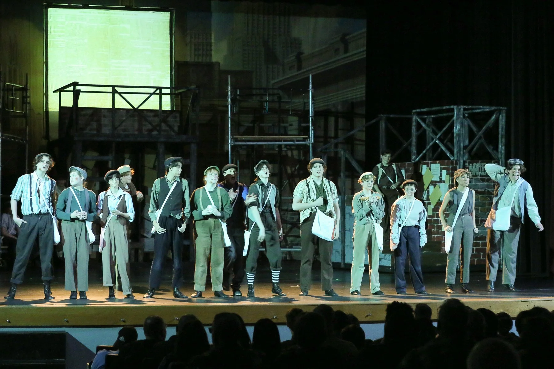 Theatrical stage with actors dressed as poor workers or students, standing in a line, performing a scene. The background features urban scenery with scaffolding, a brick wall, and a large illuminated backdrop.