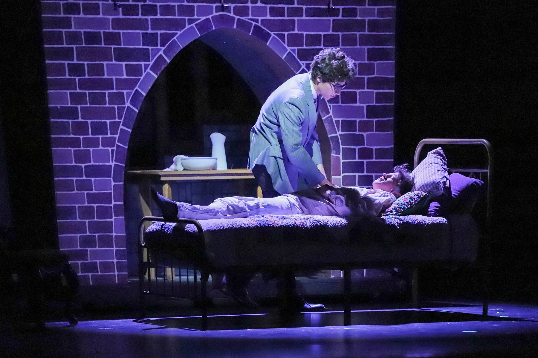 Stage scene of two actors in a theater performance. One actor is lying in a hospital bed, looking up, while the other actor stands next to them, holding their hand. The background features a brick wall with an archway.