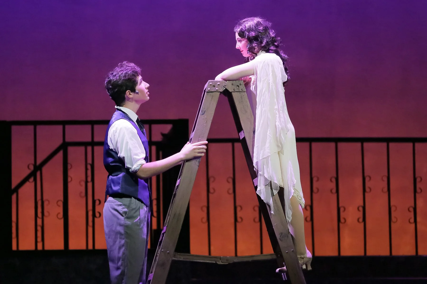 A man and a woman on stage, with the woman standing on a ladder and leaning on the top rung, looking at the man. The stage background is a gradient purple to orange, with a black iron fence silhouette.