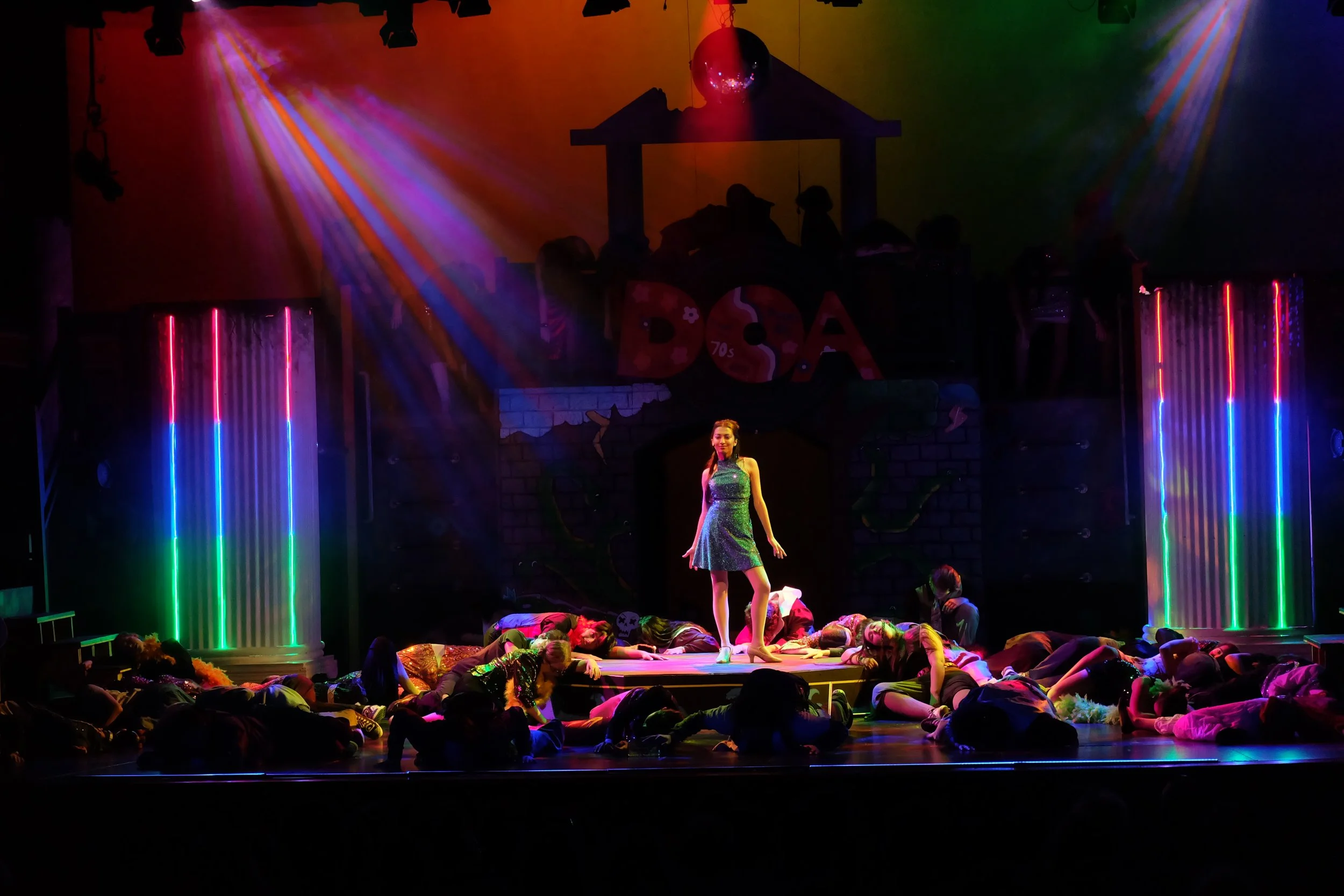 A theatrical stage performance with a woman standing in the center wearing a shiny dress, surrounded by children lying on the stage, with colorful lights and a backdrop of a house and rainbow-colored columns.