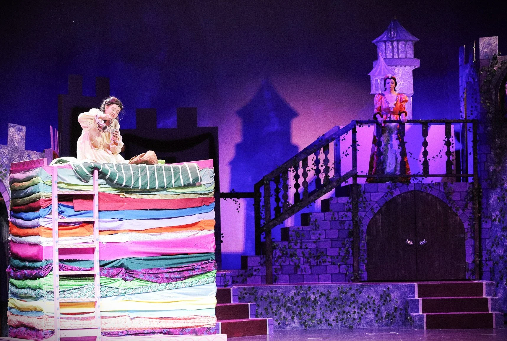 Theatrical stage scene depicting two women in period costumes, one sitting on a bed made of colorful folded blankets, and the other standing on a stone balcony with a castle-like backdrop, purple and blue lighting enhances the fantasy setting.