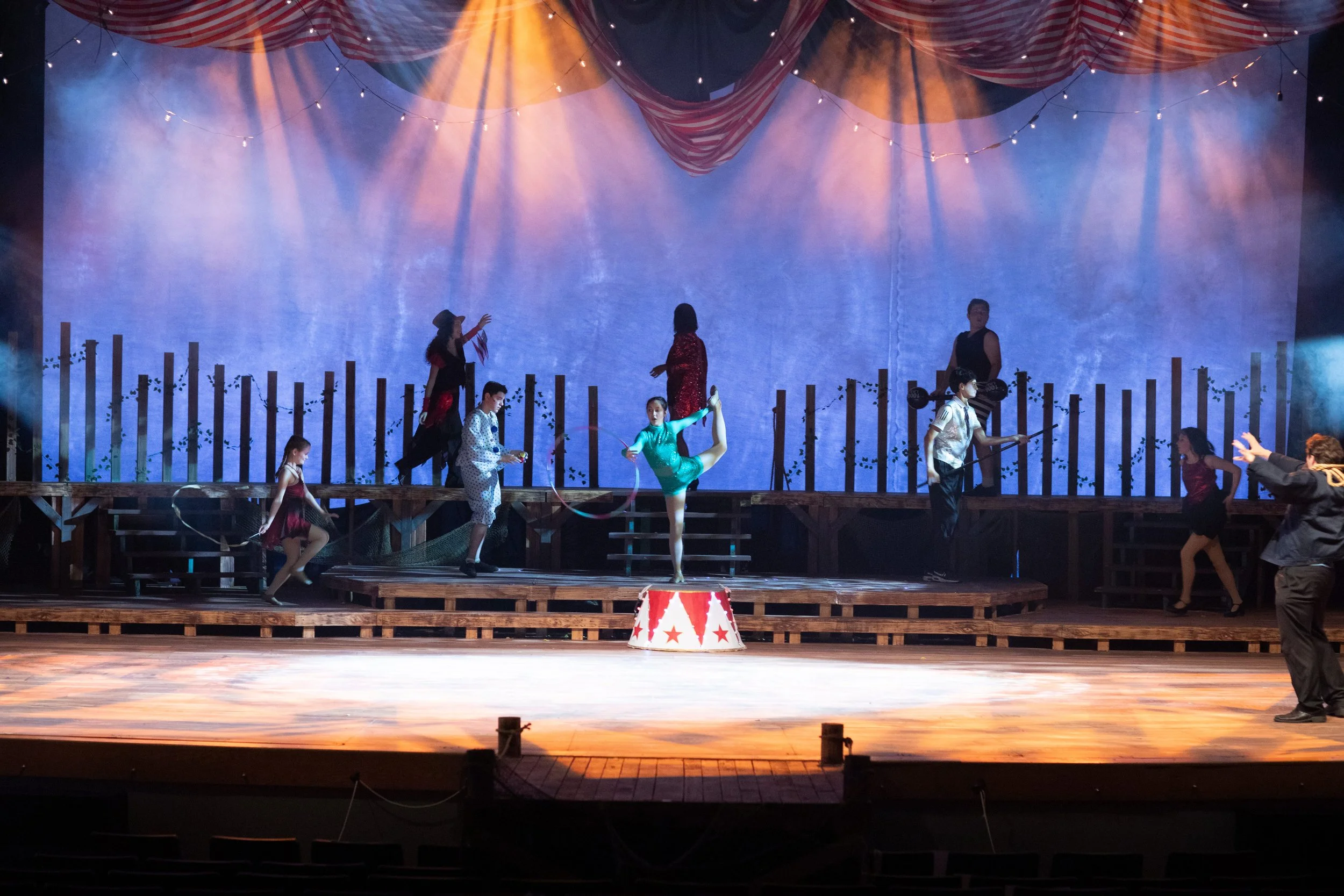 A circus stage with performers, including a woman in a green costume doing a split, other performers in colorful costumes, and a man in a striped vest. There is a large, draped red and white striped circus tent overhead with string lights. The stage 