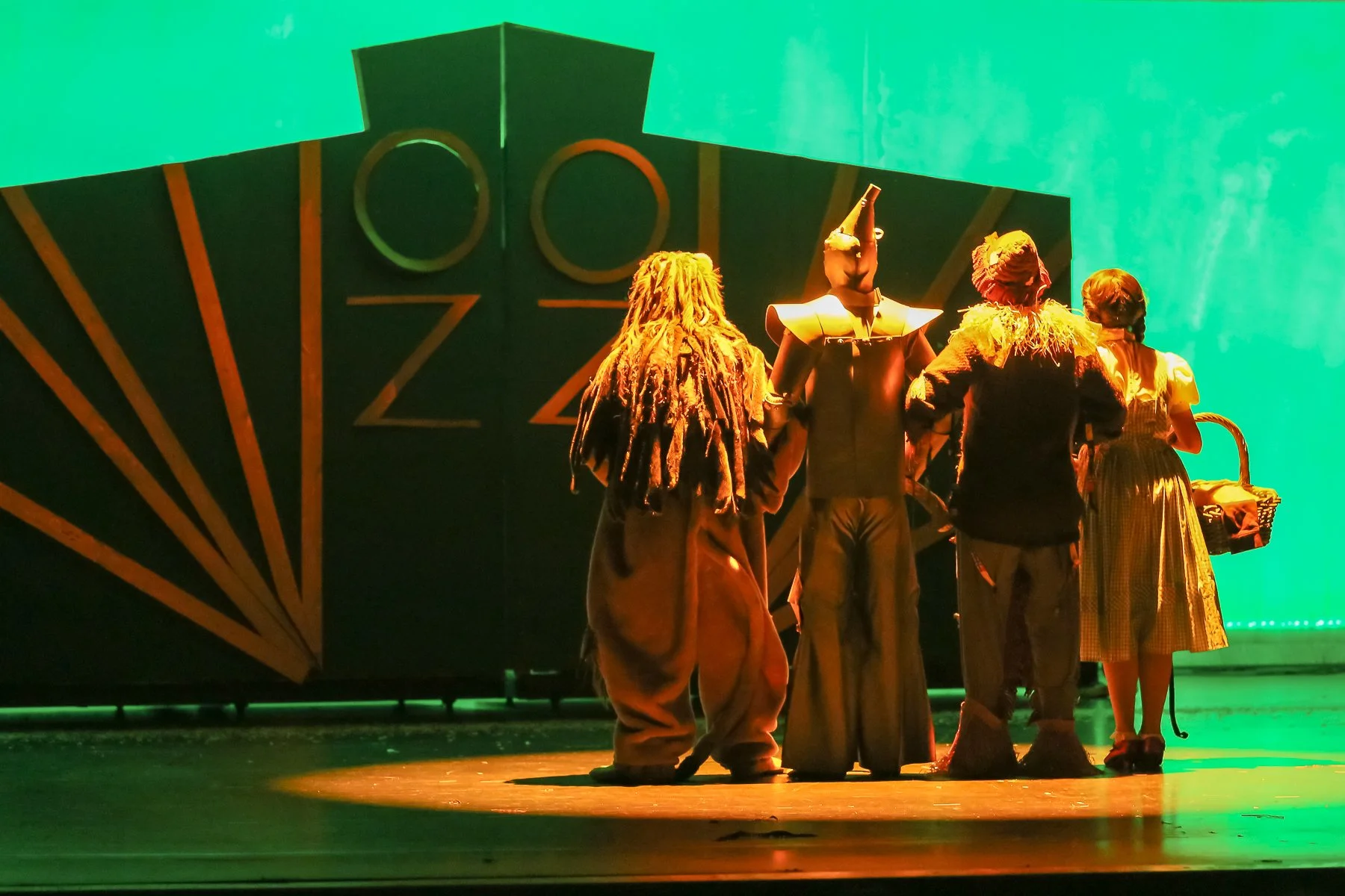Stage performance of characters dressed in costumes, including a character with a tall hat and others with elaborate dresses, against a green backdrop with a black structure with circular and triangular design elements.