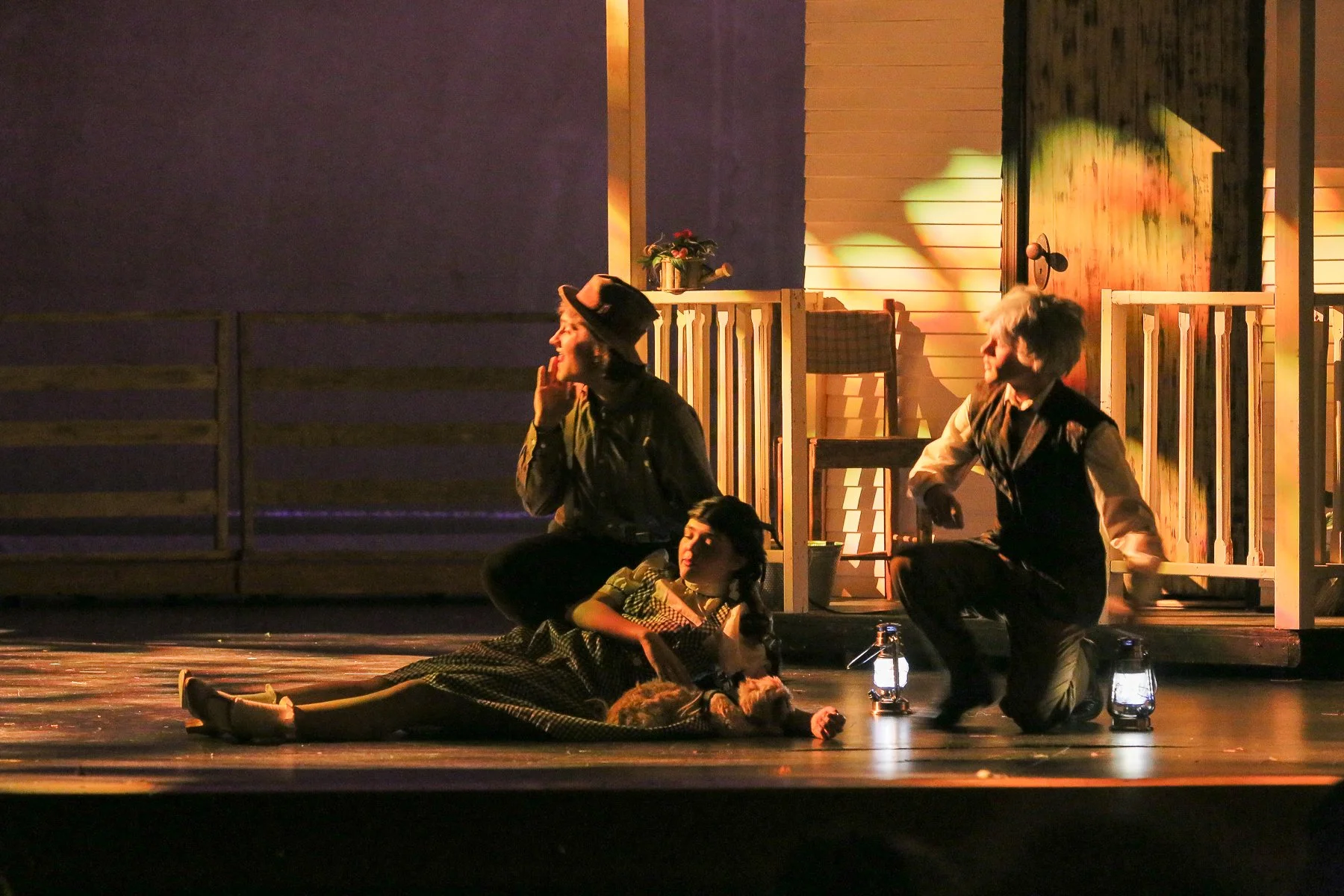 The image shows a stage scene with actors in period costumes, with a wooden porch and flowers in the background. One woman in a hat and dark dress is sitting, smiling and touching her face, while a young girl lies on the floor, looking up. A man in a