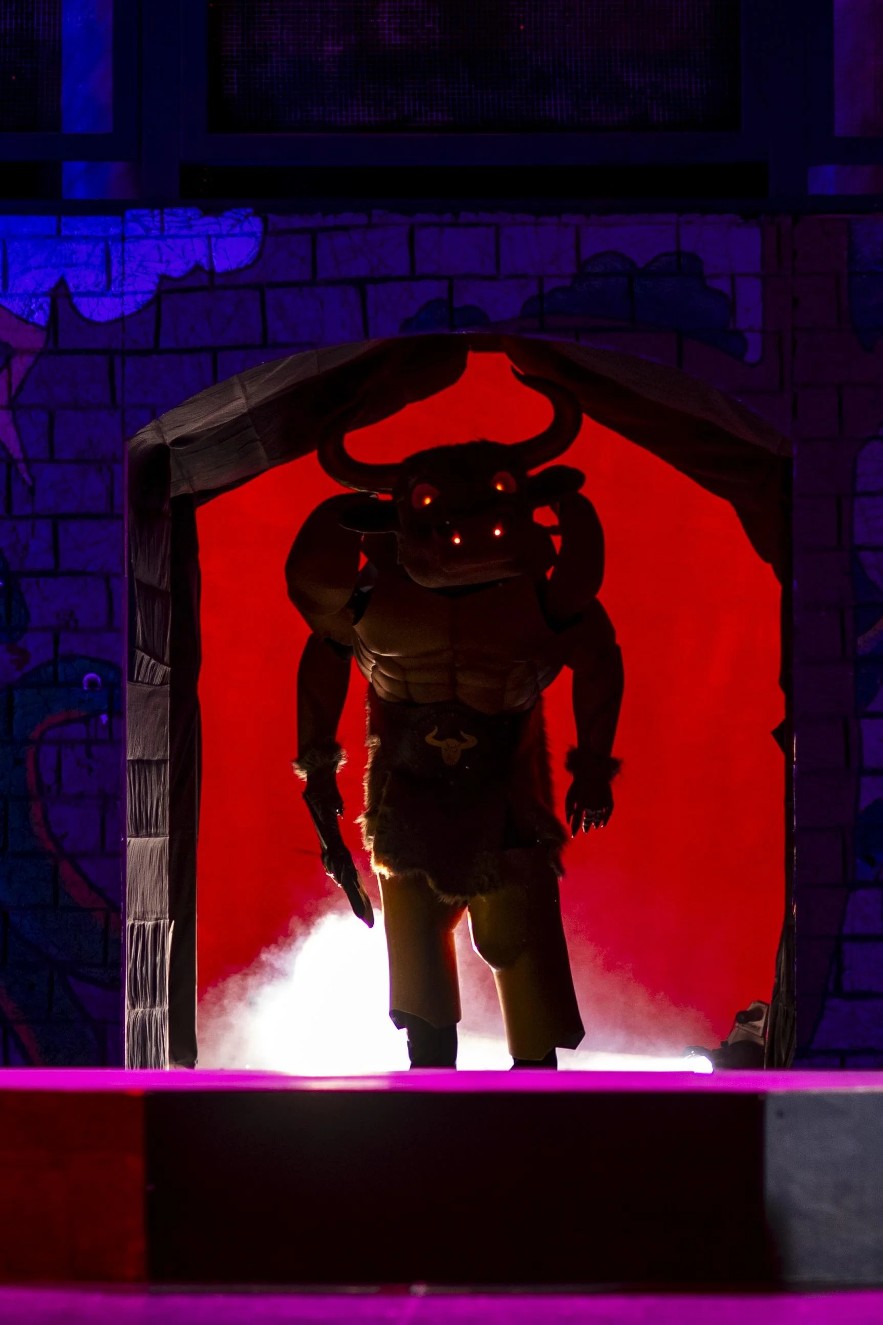 A person wearing a buffalo mask and furry costume, walking out from a red-lit doorway with purple and blue lighting on the brick wall background.