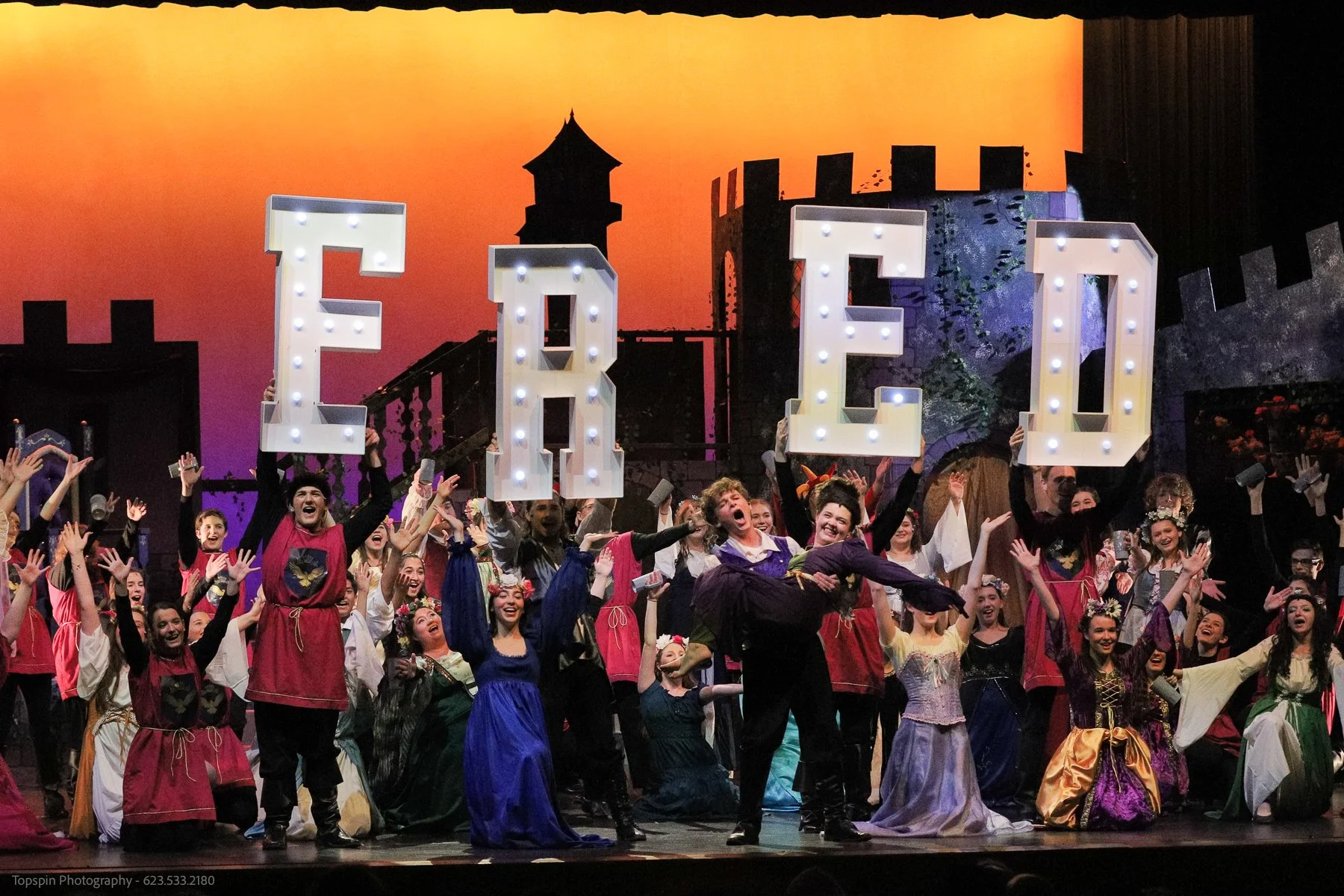 A large group of people in costumes on stage celebrating during a theatrical production, holding illuminated letters spelling 'EGO' with a castle silhouette and sunset backdrop in the background.