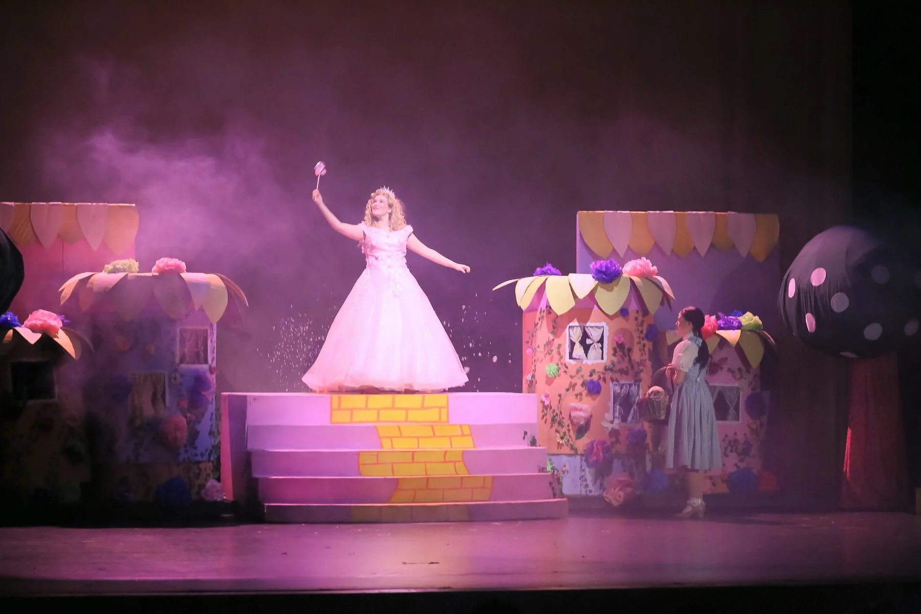 Stage scene from a theatrical performance of 'The Wizard of Oz' featuring a woman dressed as Glinda the Good Witch standing on a pink staircase, surrounded by colorful whimsical set pieces representing a fairy-tale world.